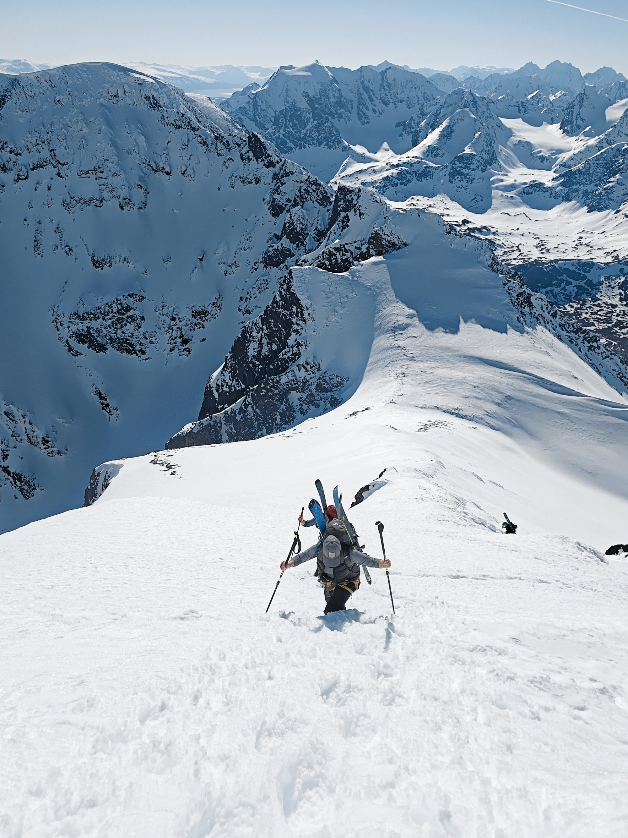 A view from behind a lone skier climbing a steep, exposed snow-covered mountain face.