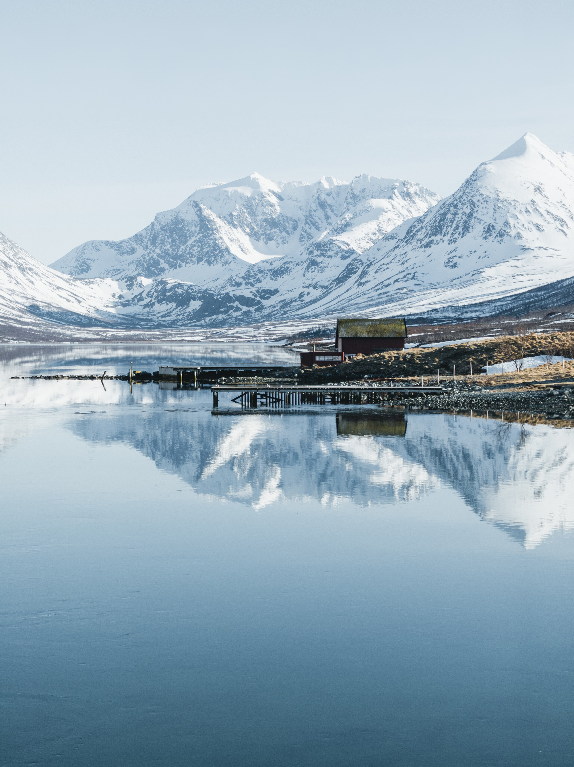 A serene landscape showing a quiet wooden shoreline hut and a small dock reflecting symmetrically into the perfectly calm, glassy surface of an Arctic sea fjord.