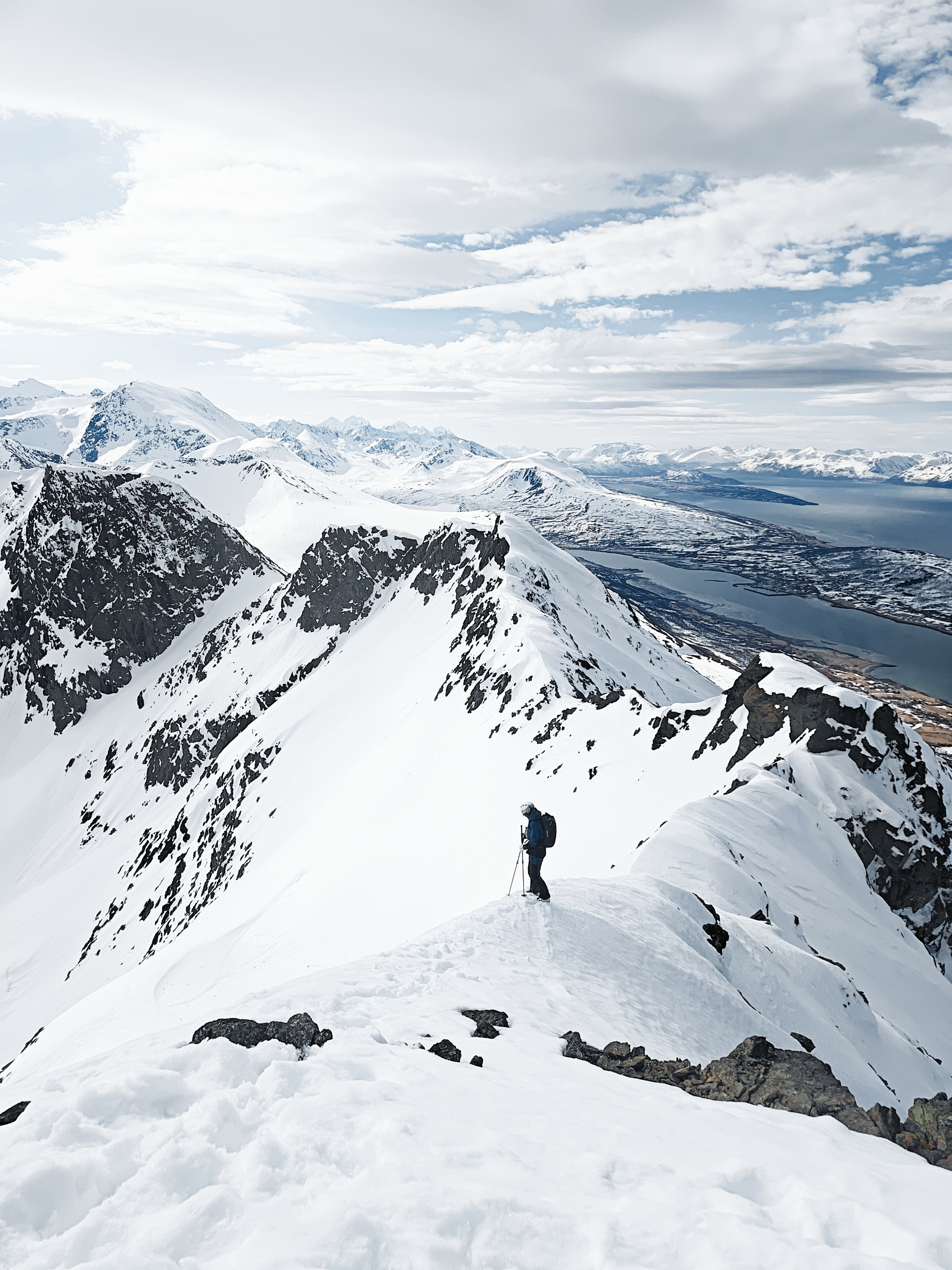 A stark, wide landscape shot focusing on a lone mountaineer carrying a backpack on a snow-packed mountain ridge line under a soft winter sky.