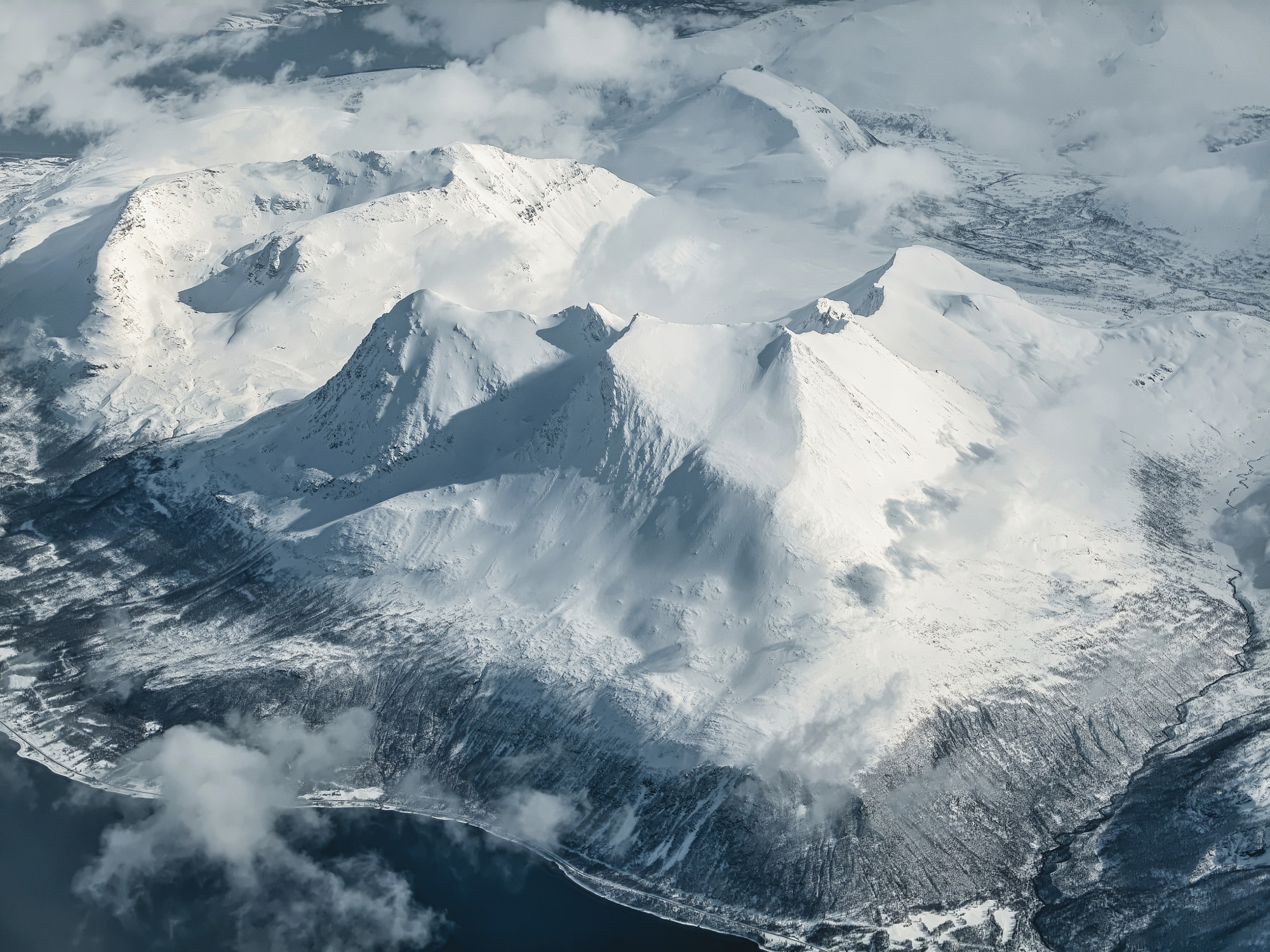 An aerial, high-altitude perspective of sharp, snow-covered triangular mountain peaks with a layer of mist and clouds.