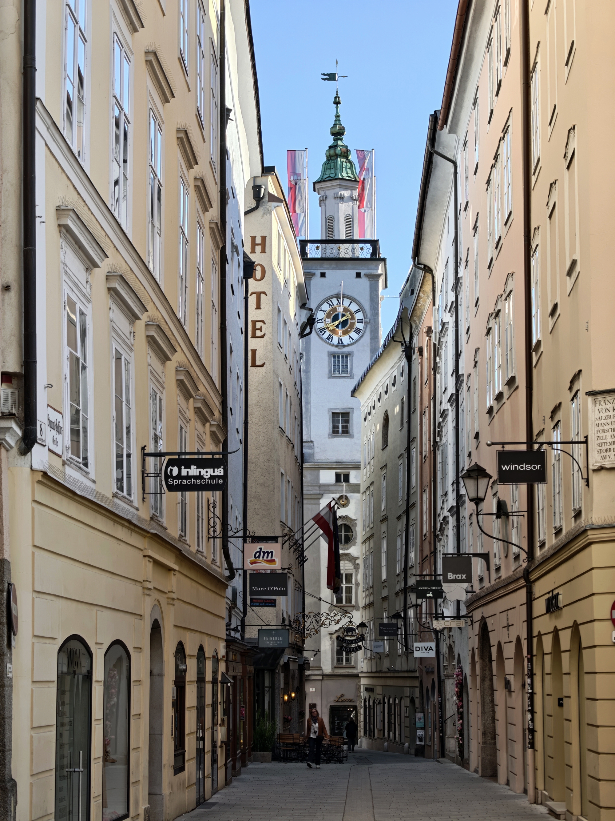 A narrow vertical composition framing a tall, historic white clock tower with a prominent green spire at the end of a cobblestone street in Salzburg's Altstadt.