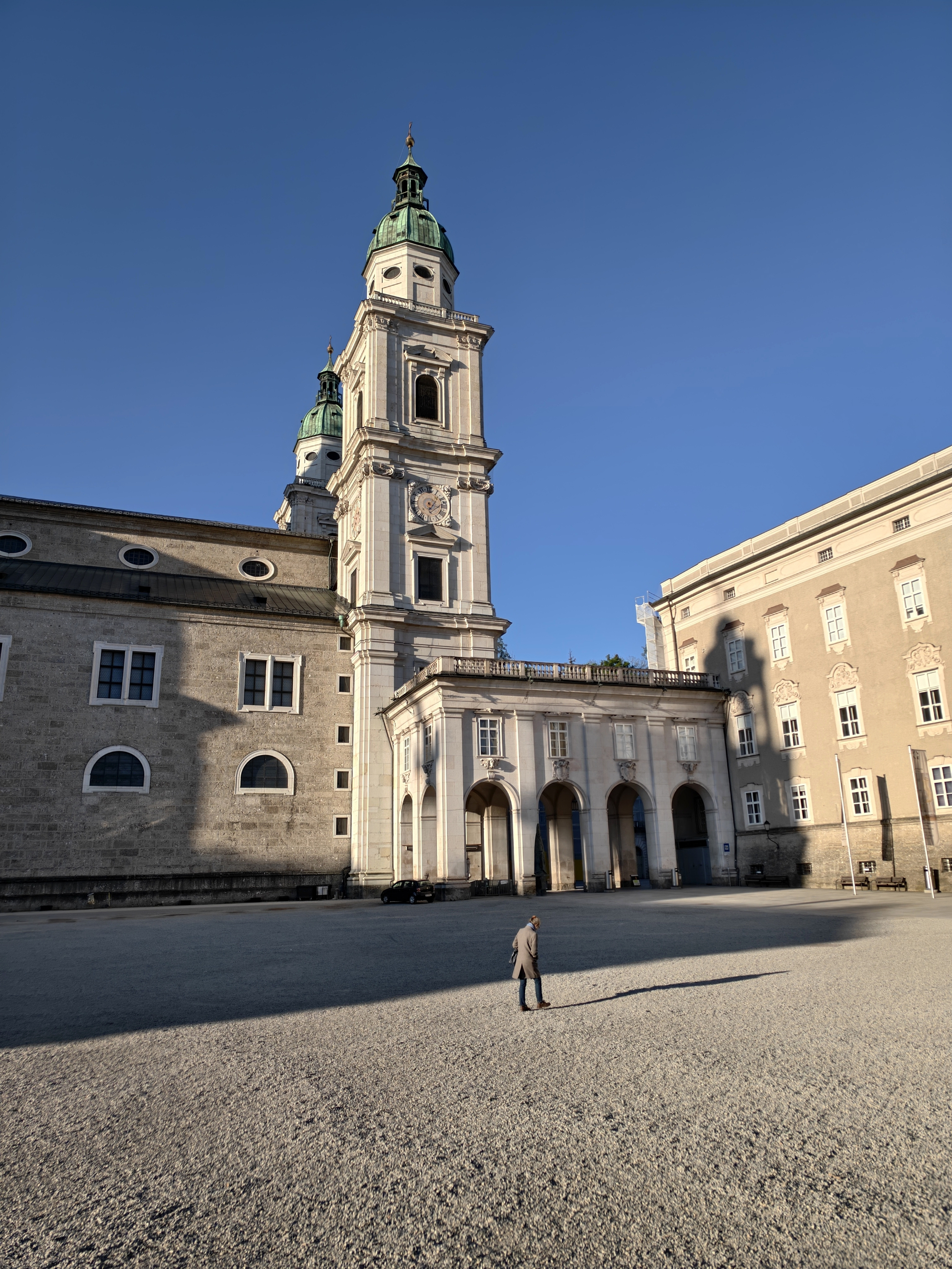A stark, geometric architectural shot showcasing a lone traveler walking across an expansive, sun-drenched stone courtyard right next to the historic Salzburg Cathedral tower.