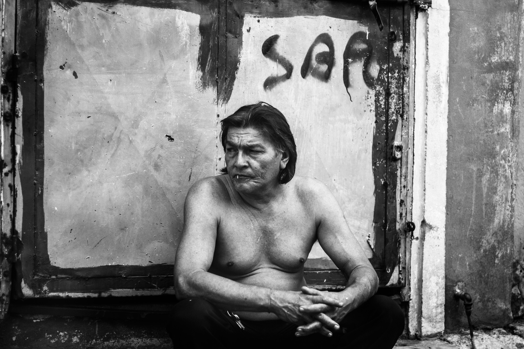 Bare-chested man sitting by a graffiti door.
