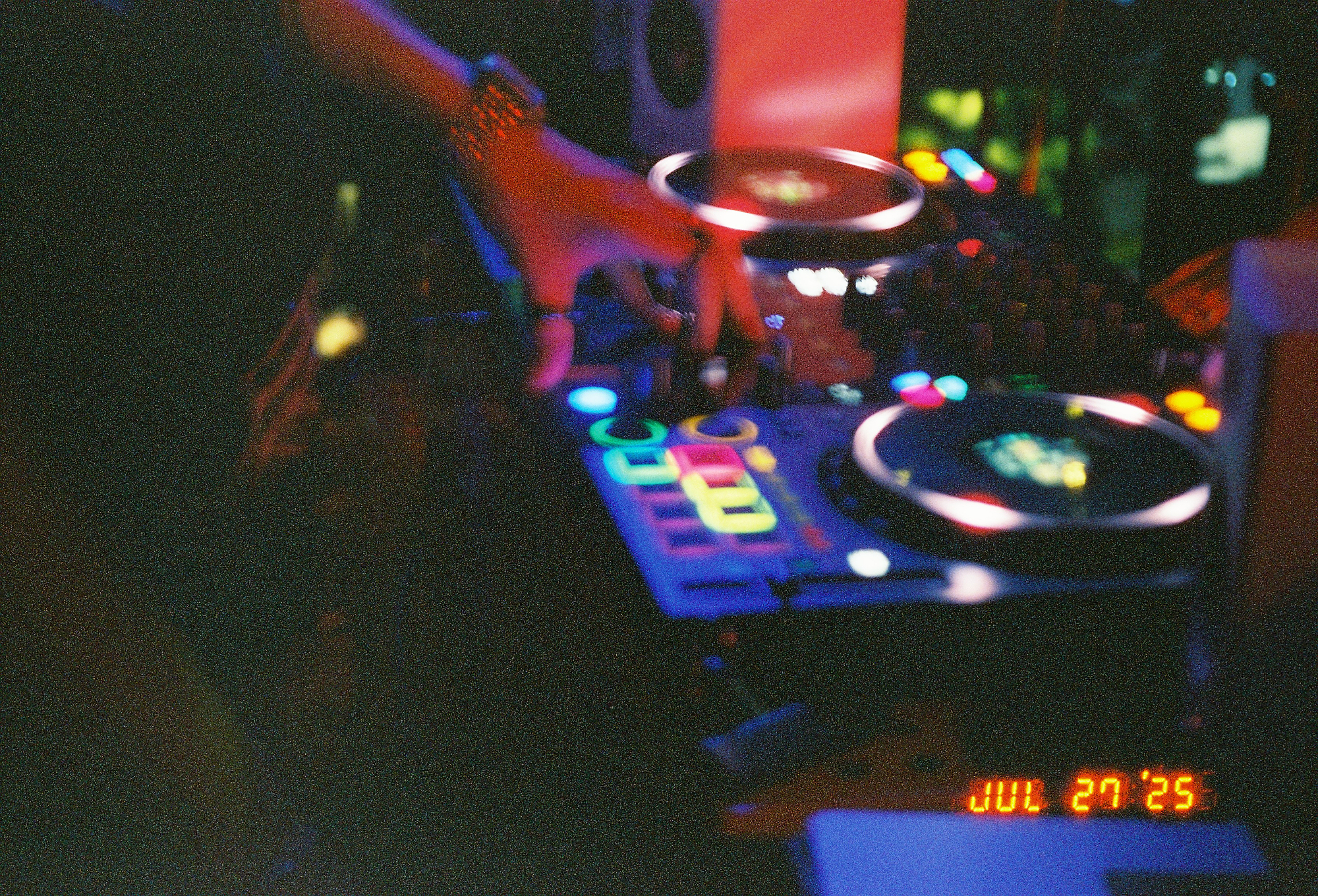 Close-up of a DJ's hand on a glowing controller, captured on grainy night photography film with a retro timestamp.