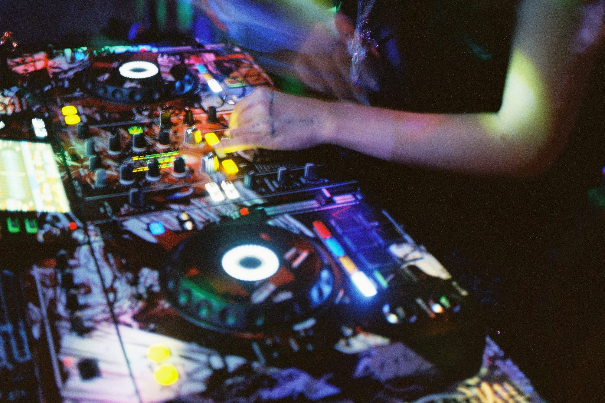 Analog music photographer shot of a DJ's hands working on an illuminated pioneer soundboard at a crowded night event.