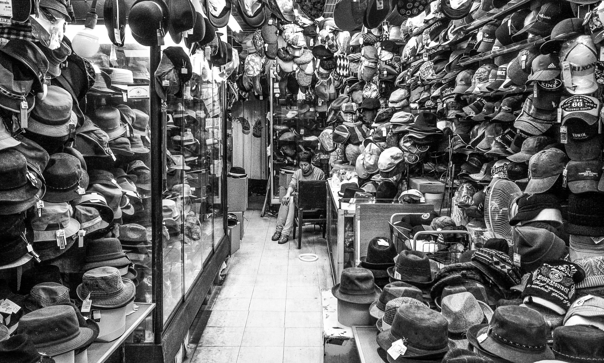 Candid black and white interior shot of an old shopkeeper sitting inside a tightly packed Japanese hat store, captured by a Japanese street photographer.