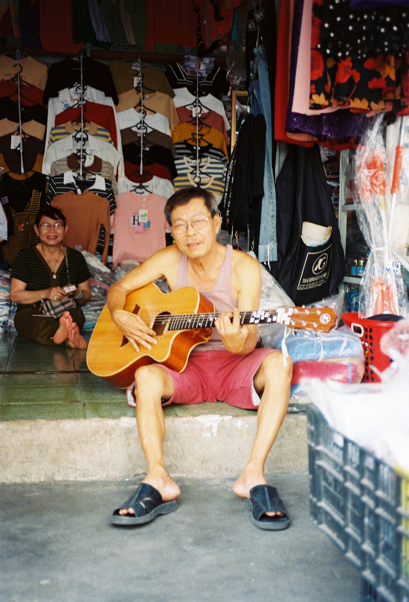 Southeast Asia street photography of an older man playing an acoustic guitar outside a local clothing market stall.