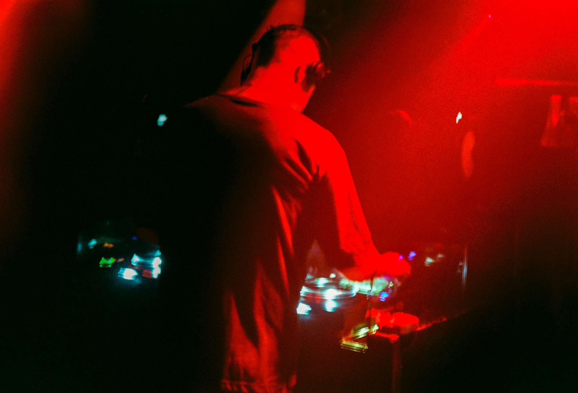Raw 35mm film photography of a DJ performing under heavy red ambient stage lighting at a night music event.