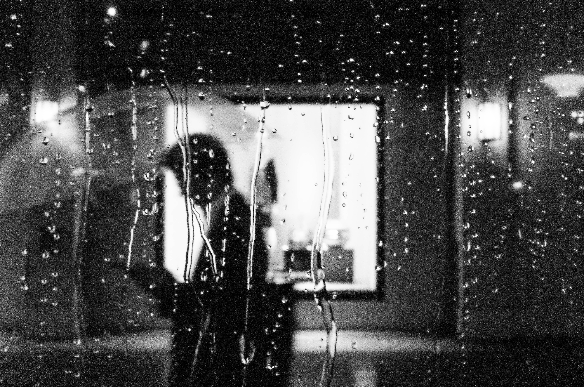 Heavy rain droplets on a glass window pane blurring a silhouetted pedestrian carrying an umbrella outside, from the Masahiro Miyagi street photography collection.