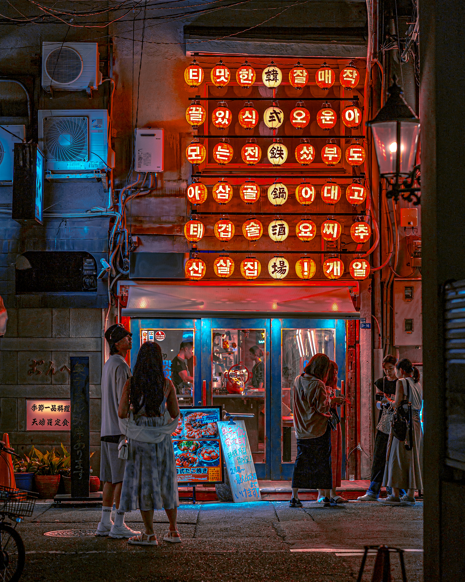 People gather outside a warmly lit restaurant at night, its facade covered in dozens of glowing red paper lanterns, highlighting the depth of 85mm street photography.