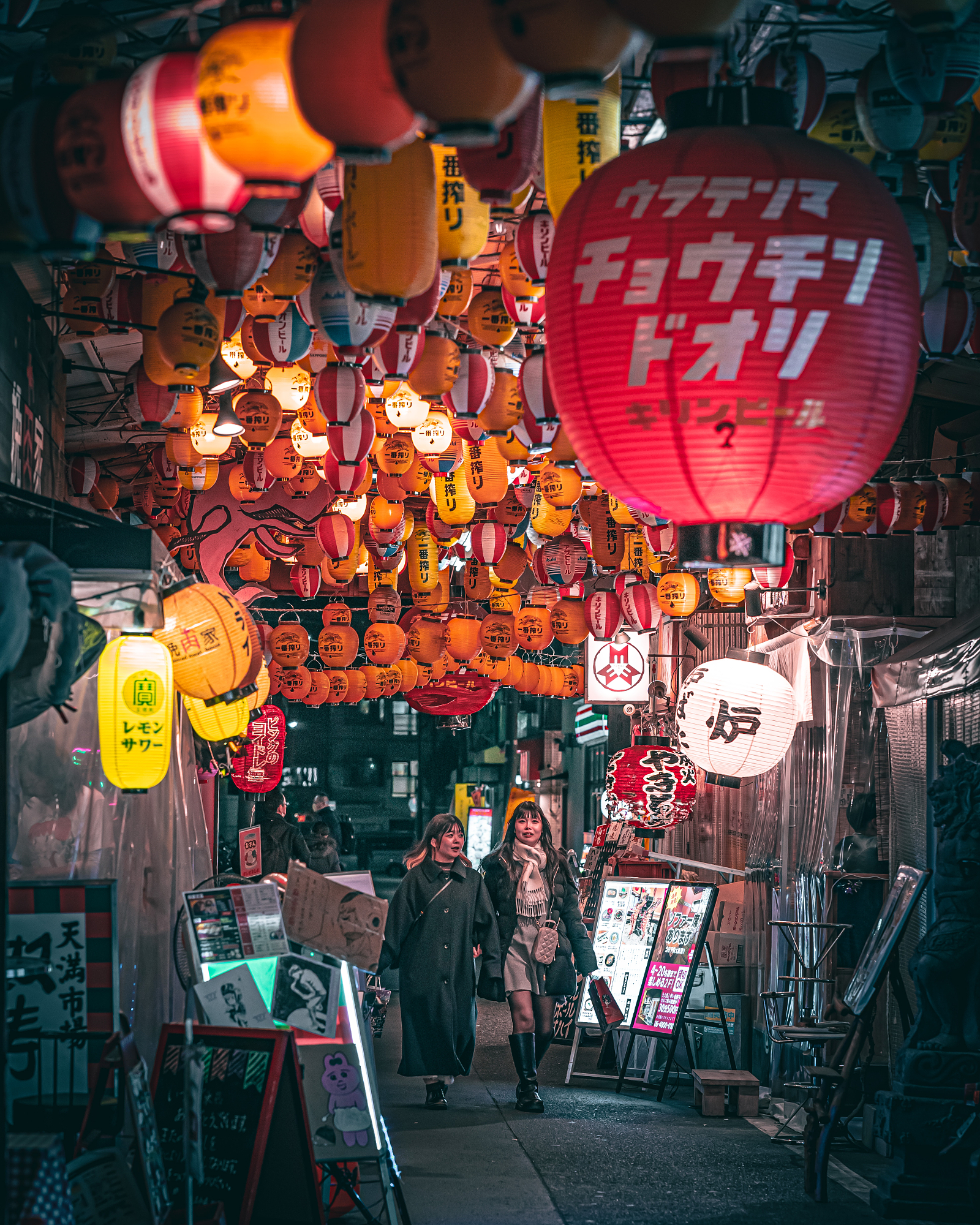 Two women walk through a dense, visually overwhelming alleyway covered ceiling-to-floor in glowing, colorful paper lanterns, utilizing the compression effect of 85mm street photography.