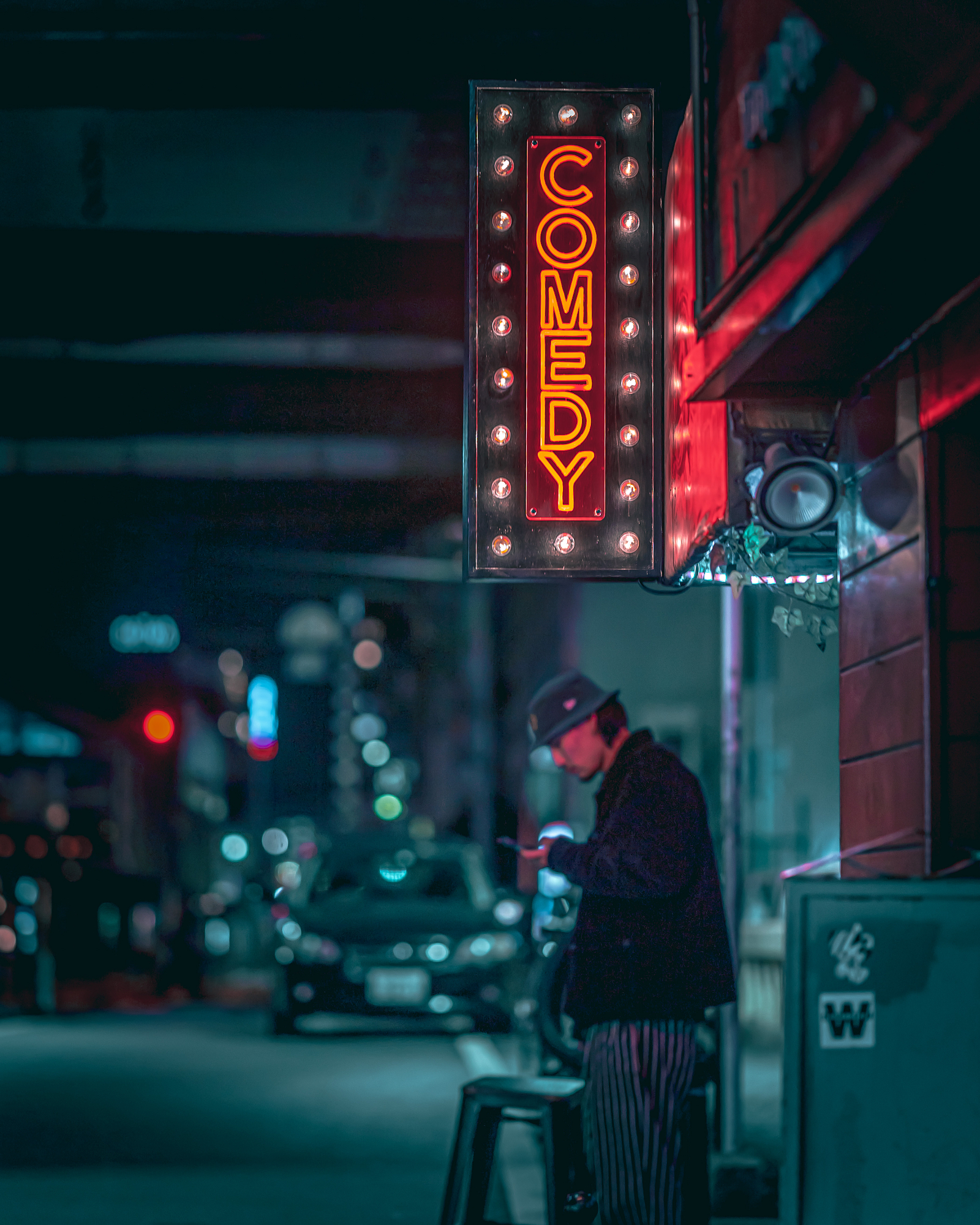 A man looking at his smartphone under a glowing red neon comedy club sign at night, capturing a moody mood in cinematic urban photography.