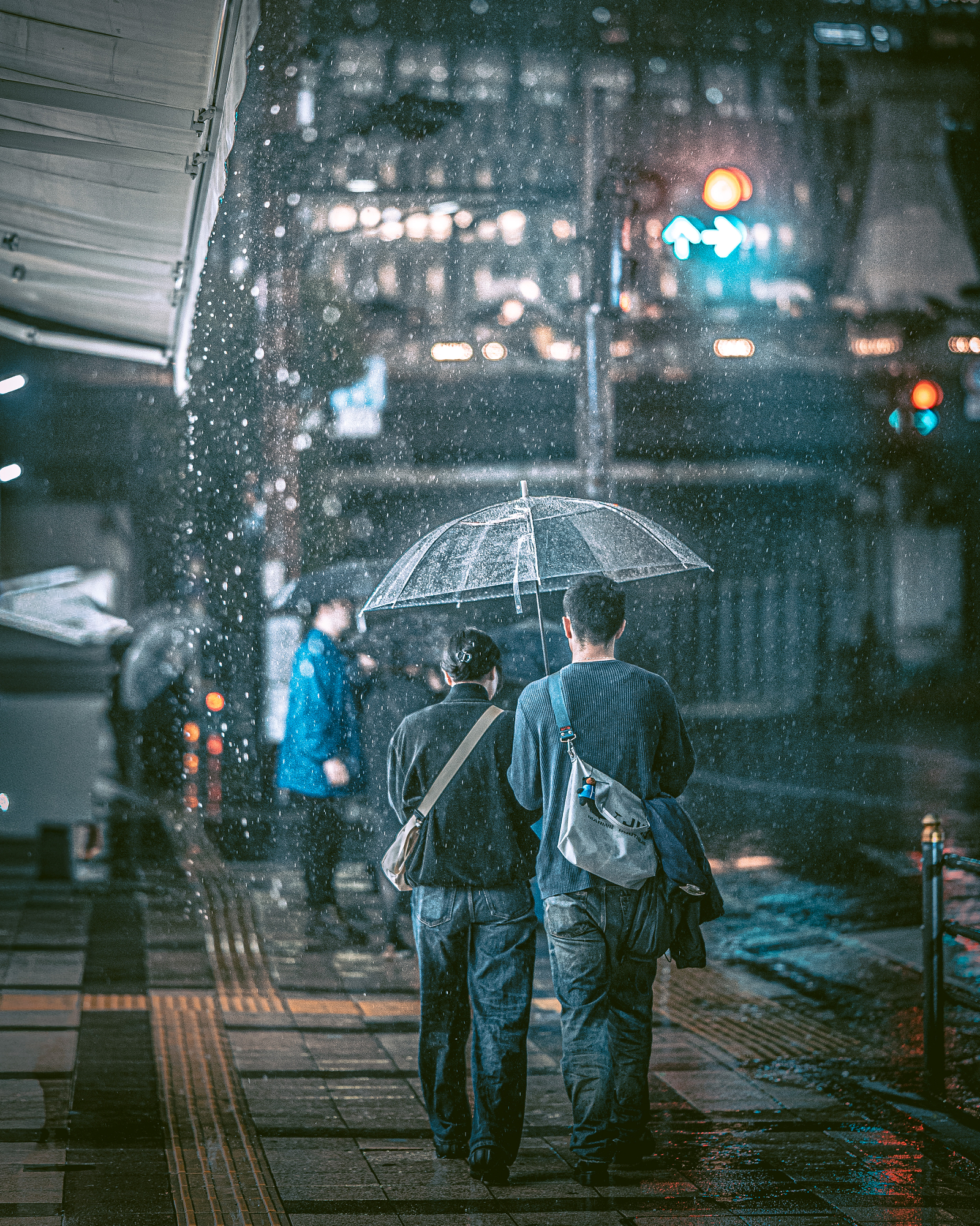 A couple walks together under a clear umbrella through heavy rain at a city intersection at night, captured beautifully by a Japanese street photographer.