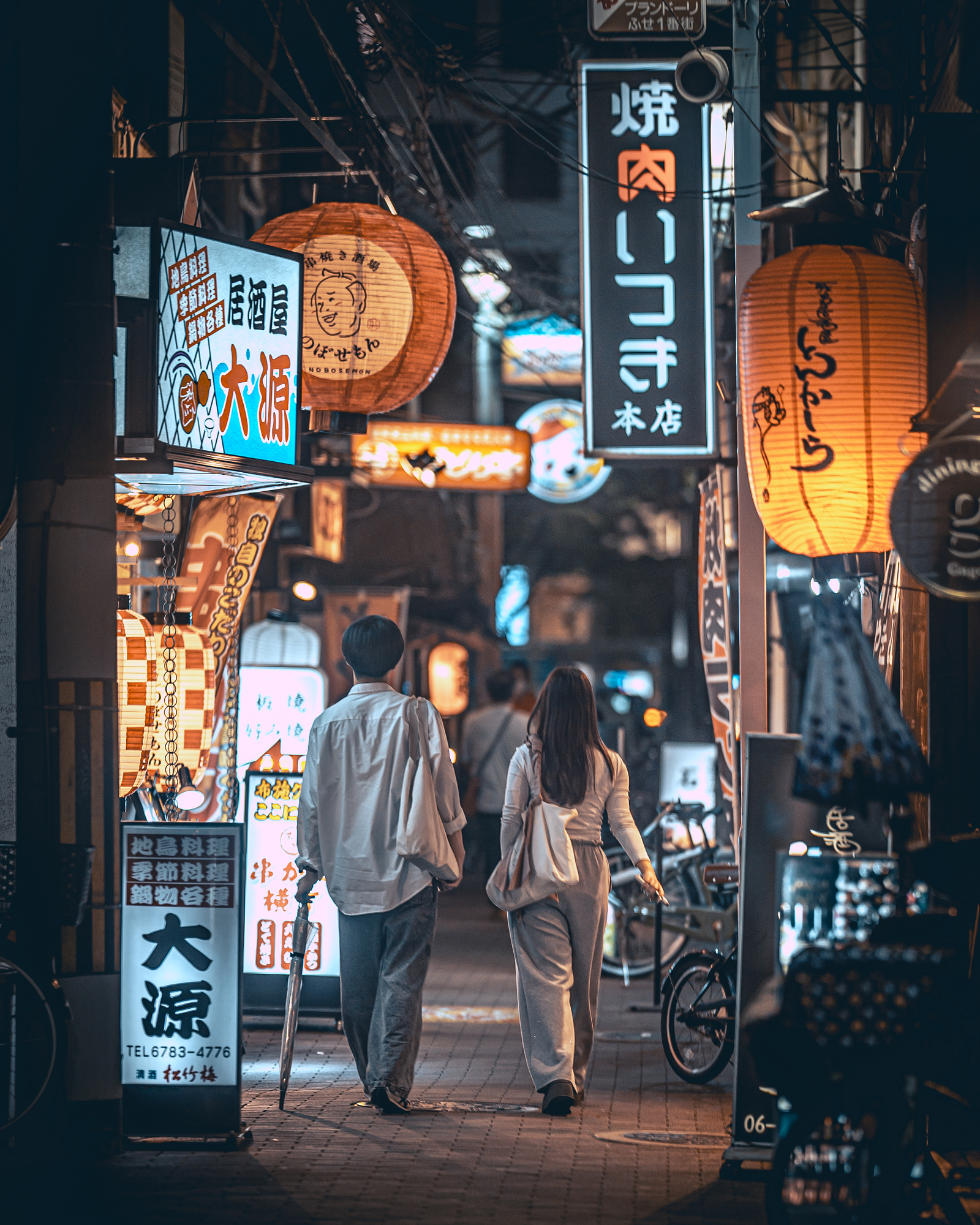 A couple walks away down a narrow, illuminated alley lined with glowing lanterns and restaurant signs, a signature composition by this Japanese street photographer.