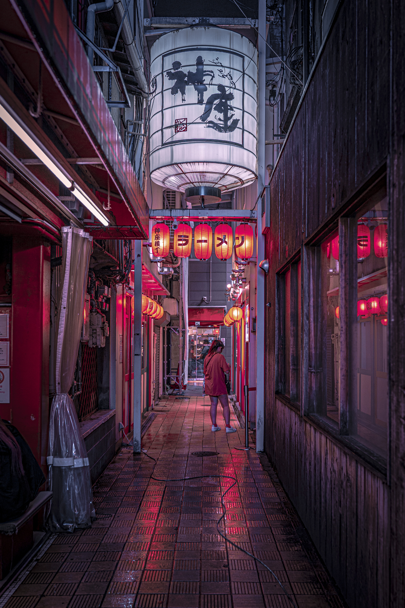 A woman in a pink shirt stands in a narrow, moody alleyway bathed in vibrant red light from hanging lanterns, a stunning example of Tokyo street photos.