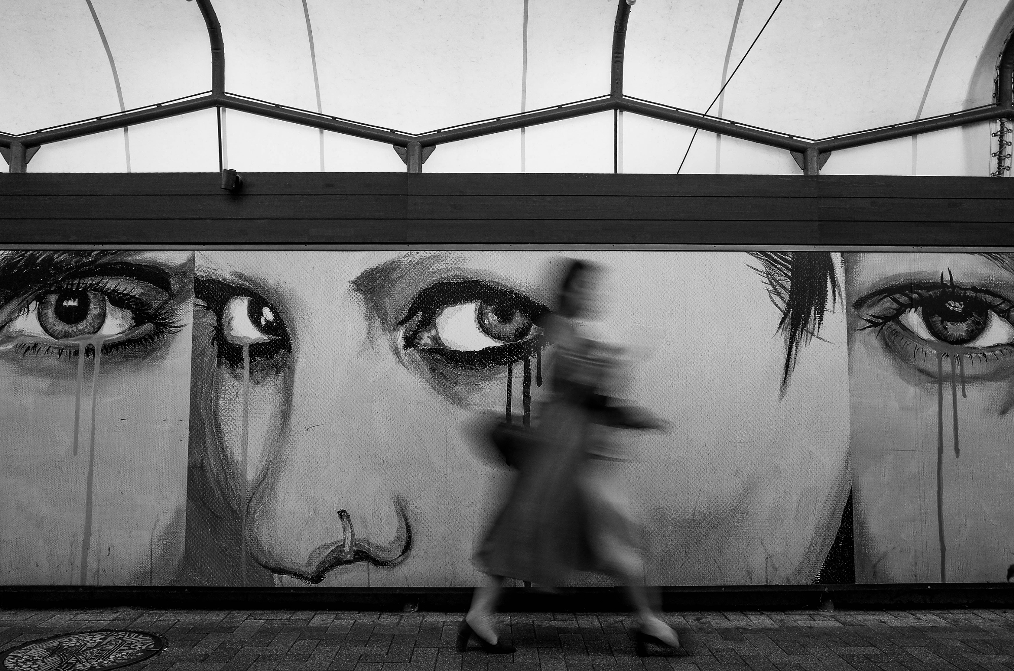 Motion blur of a person walking past a massive black and white street mural featuring detailed eyes crying, taken by a Japanese street photographer.