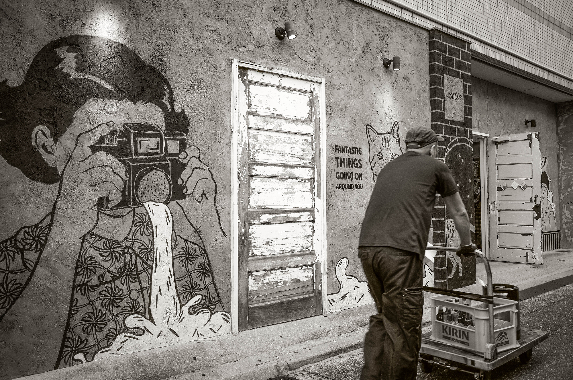 Monochrome image of a worker pushing a cart with a Kirin beer crate past a large street wall mural of a girl holding a camera, displaying unique Tokyo street photos.