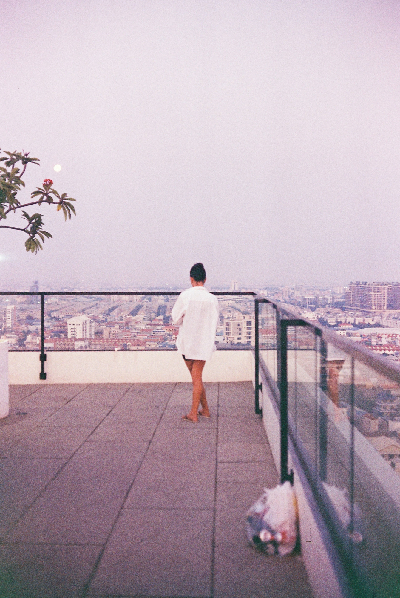 A woman in a white shirt looking out over the city skyline at sunrise from a rooftop balcony in Phnom Penh, on grainy 35mm film.