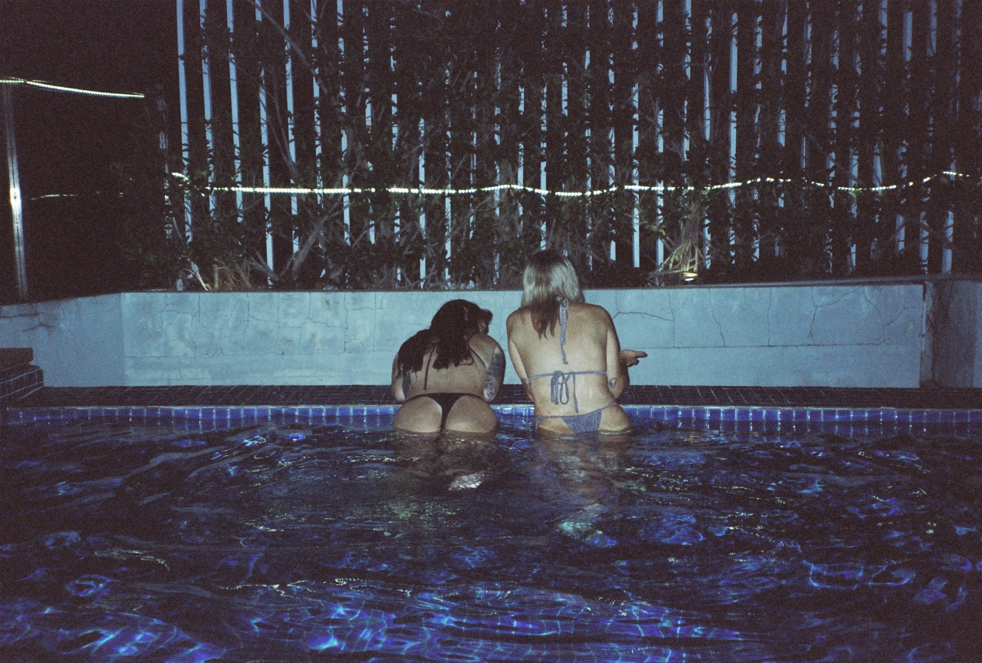 Analog music photographer style night snapshot of two women relaxing in an outdoor swimming pool against a wooden fence.