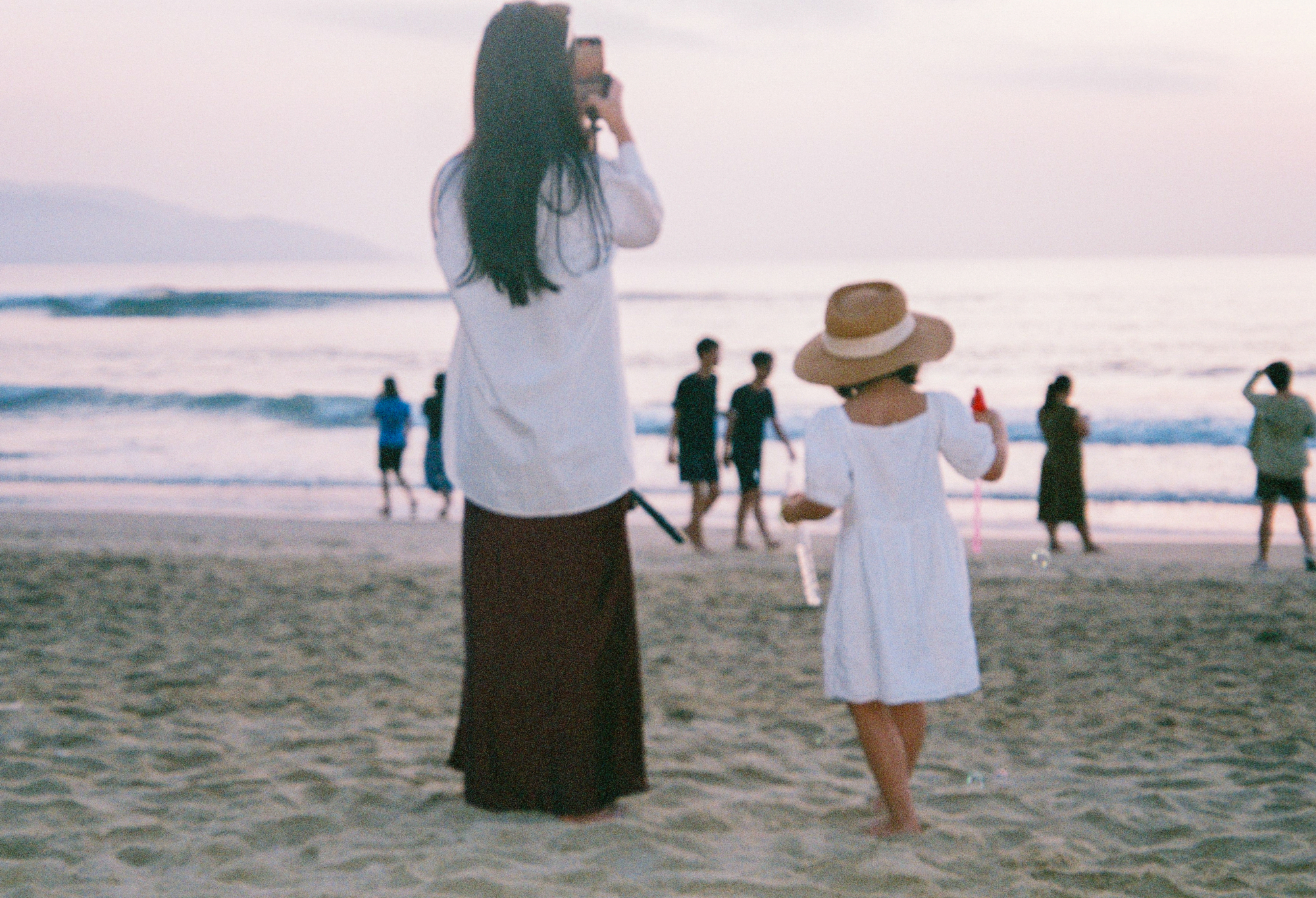 Nostalgic travel photography on 35mm film showing a mother taking a picture of her child on a sandy beach during sunset.