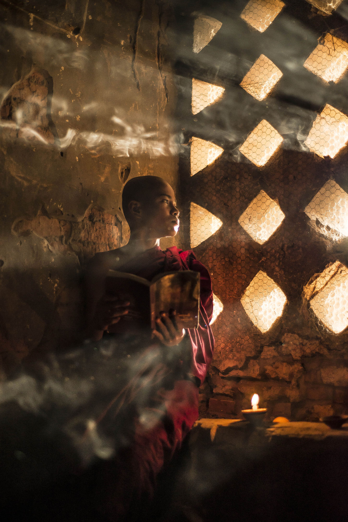 A young monk reading a book next to a patterned lattice window, creating a dramatic rim light portrait with soft light.