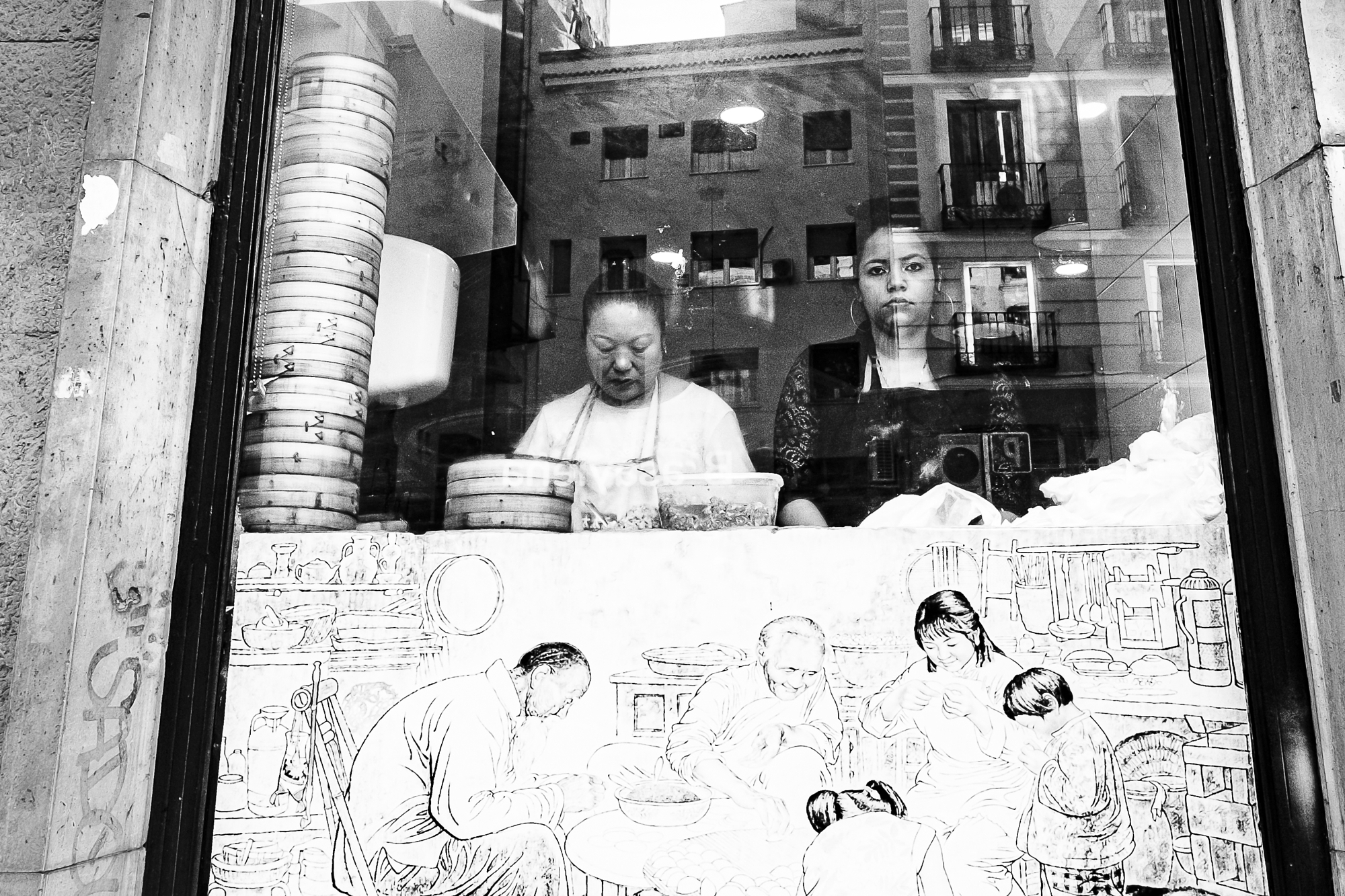 Black and white street photography of immigrant women making gyozas behind a glass window with city reflections.
