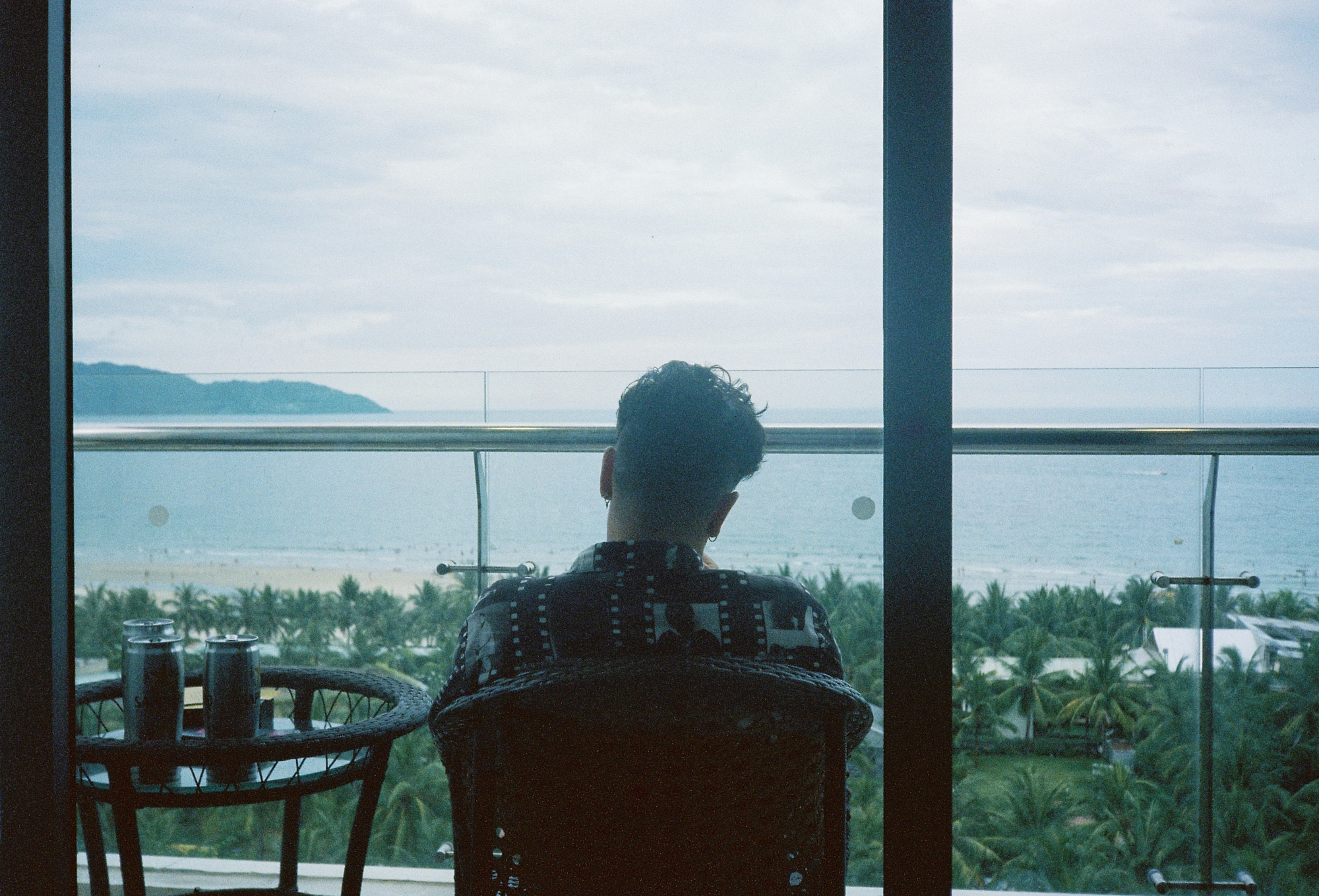 A person seen from behind sitting on a high-rise balcony looking out over the ocean beach and palm trees, captured on analog film.