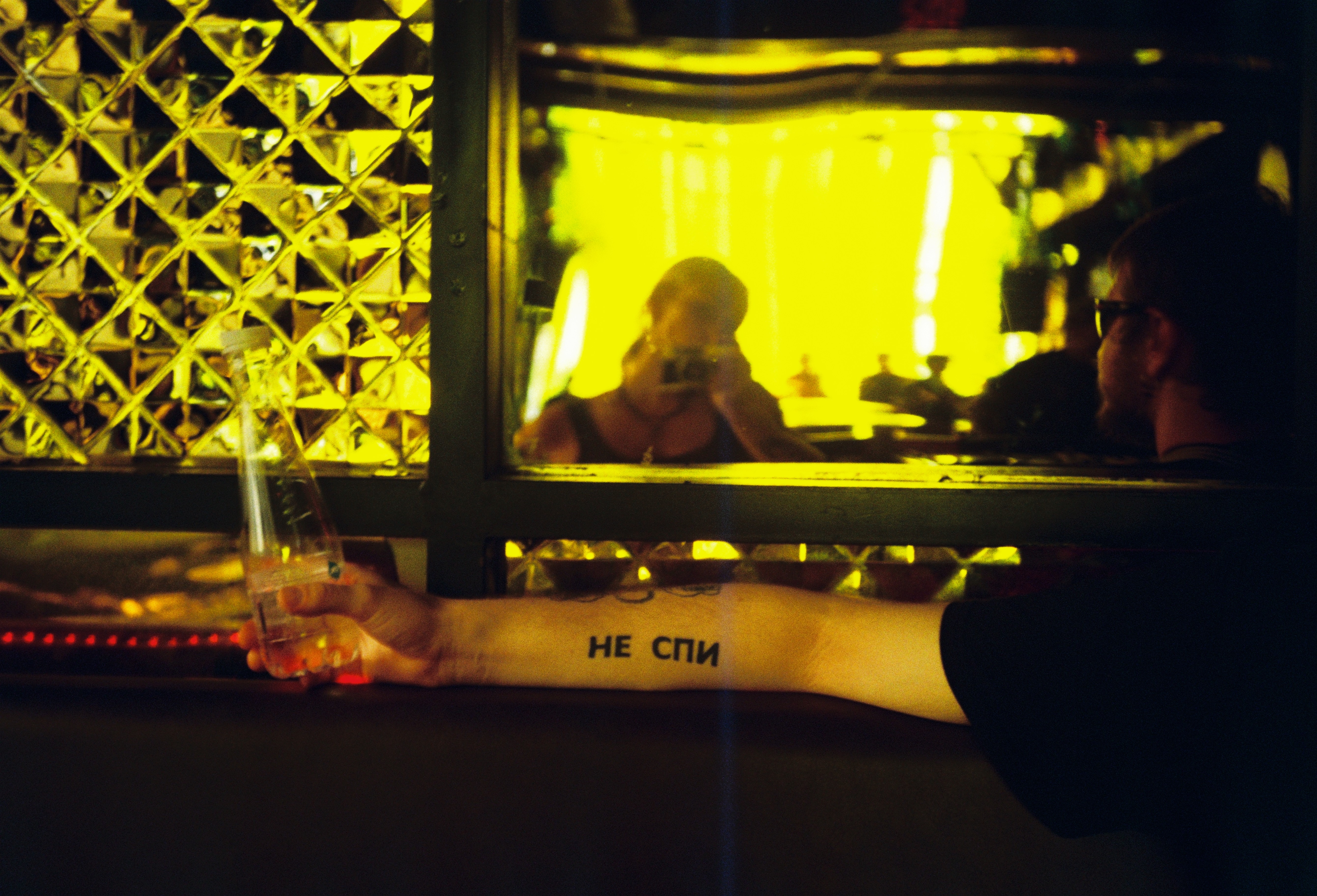 Katerina Shishkina photography capturing a person's tattooed arm reading "?? ???" at a night event under vibrant yellow neon bar lighting.