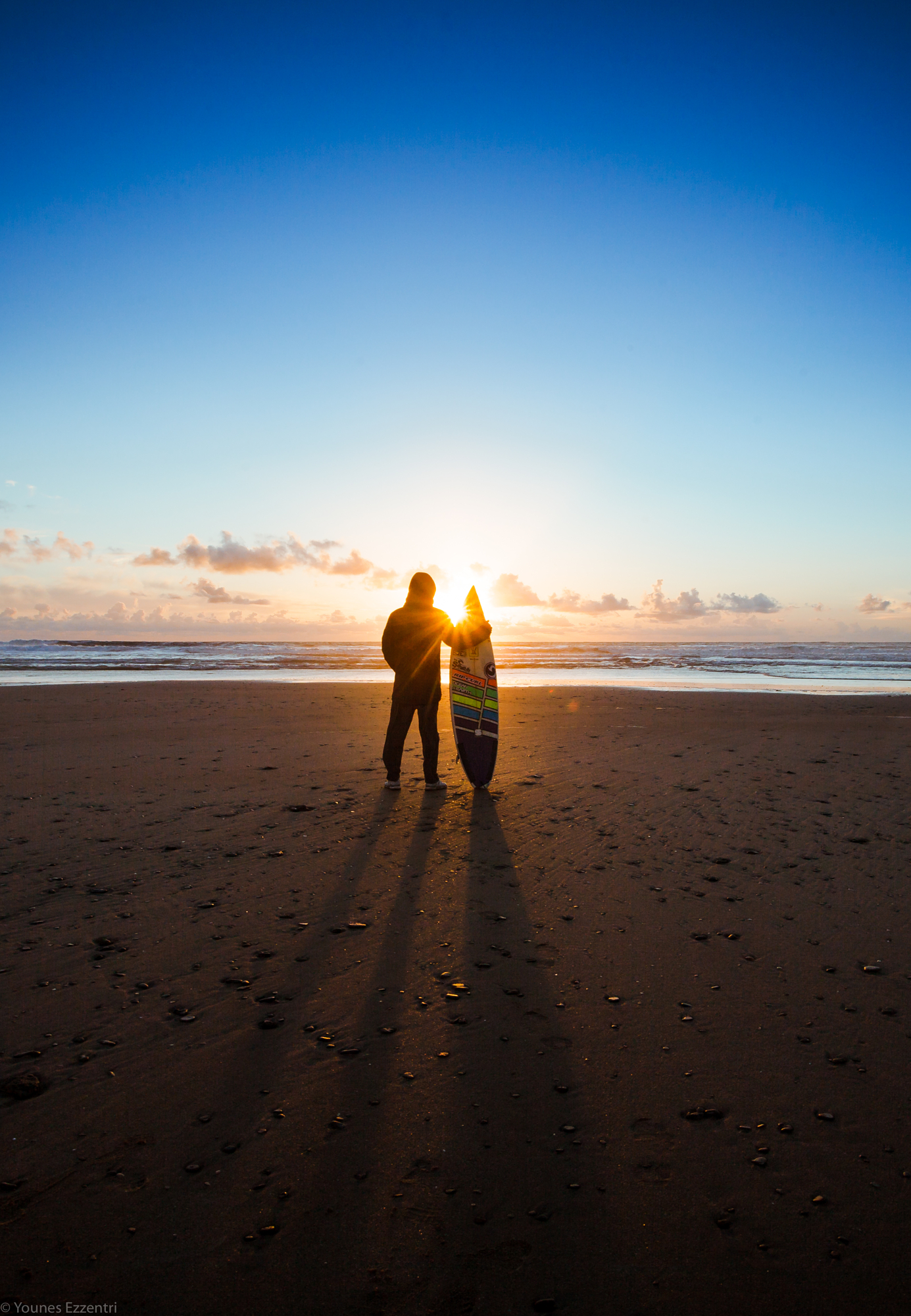 A surfer silhouette holding a surfboard on a wide beach at sunset, showing the mastery of golden hour composition in Younes Ezzentri landscape photography.