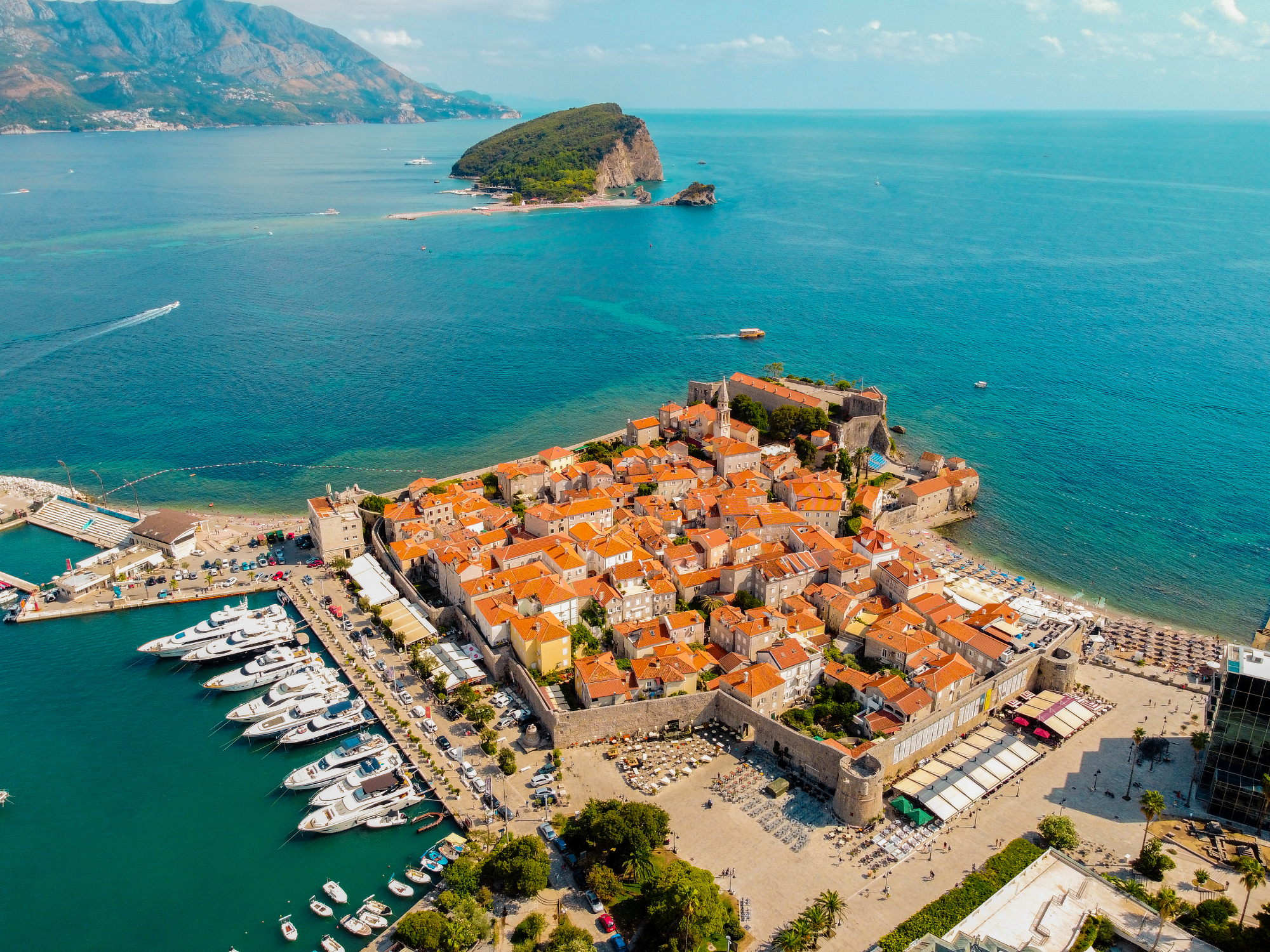 Aerial view of Budva Old Town framed by vibrant blue sea and mountains, illustrating midday photography tips for capturing landscape color while shooting in direct sunlight.