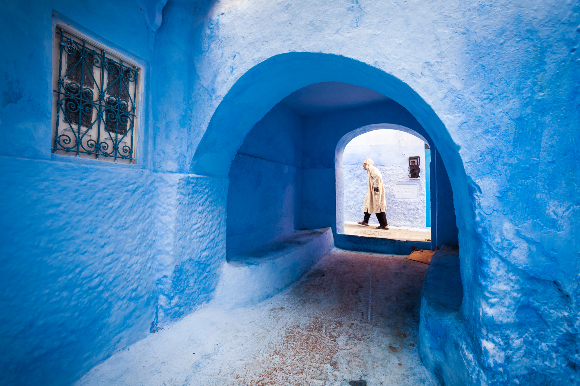 A local man in a traditional djellaba walking through a vibrant blue archway alley in Chefchaouen, showcasing unique composition in Younes Ezzentri landscape photography.