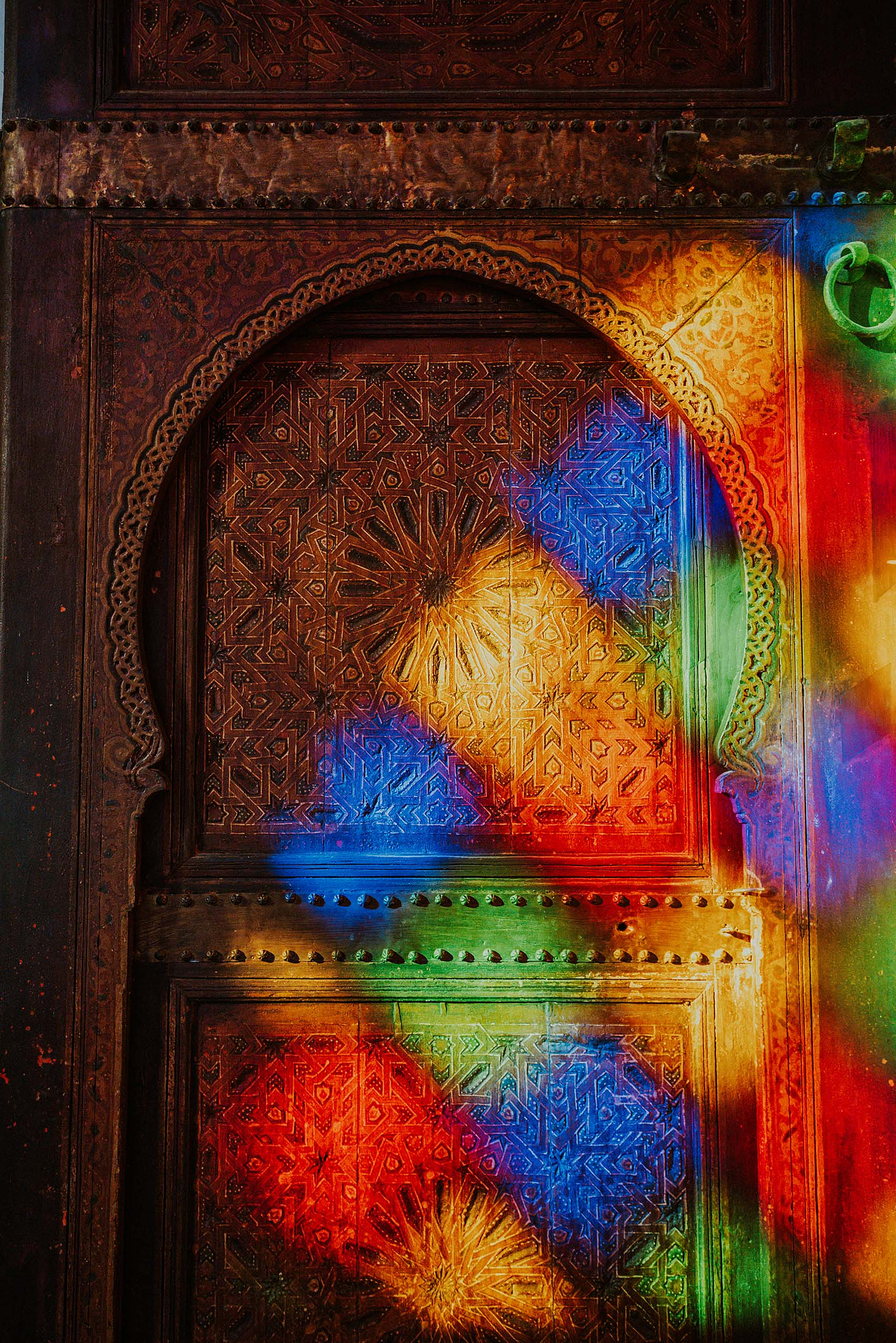 Multi-colored stained glass light reflecting vivid rainbow patterns onto an intricately carved wooden arch door, demonstrating the vibrant artistic style of Younes Ezzentri.