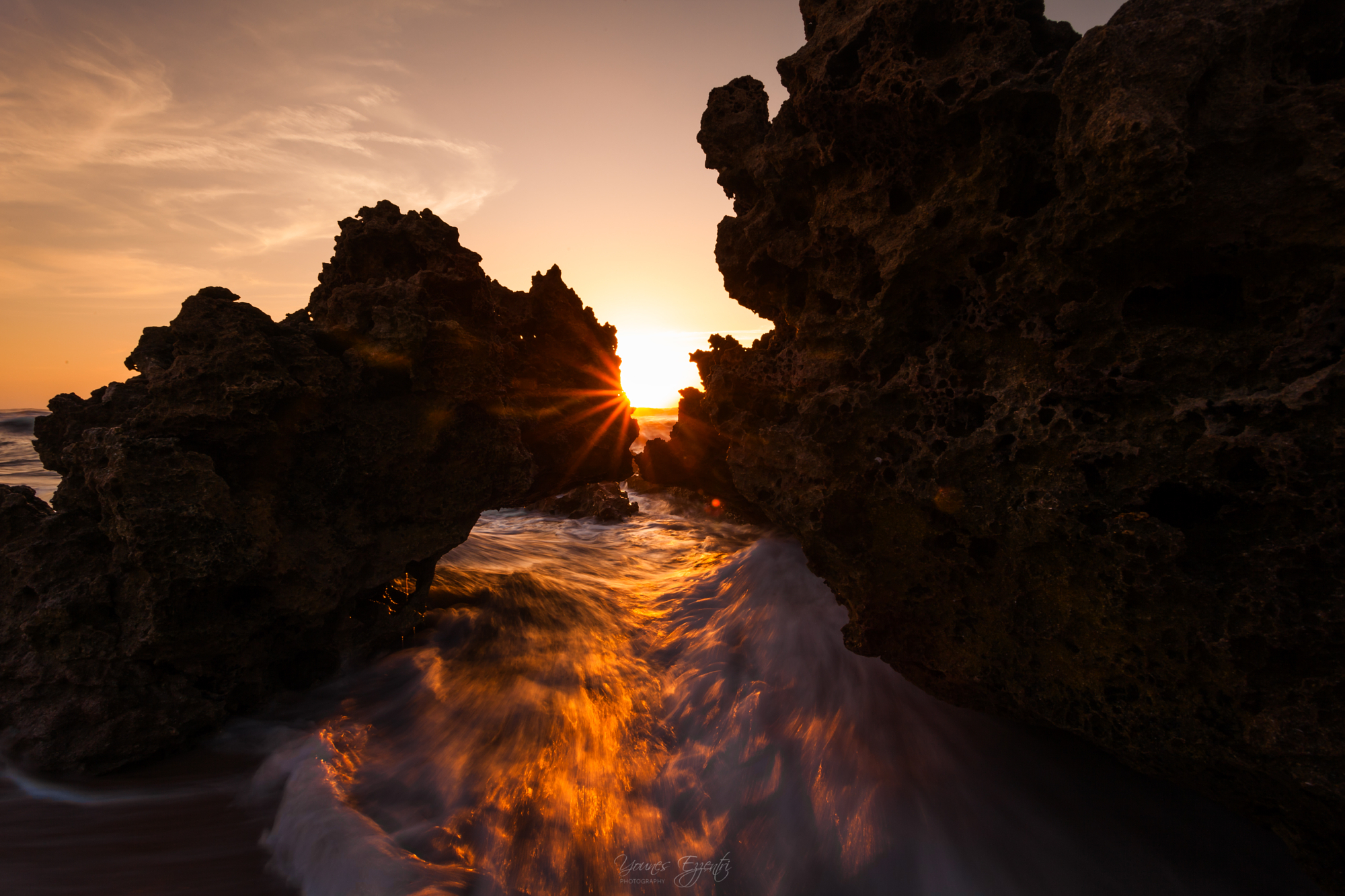 A brilliant golden hour sunburst breaking through dark coastal rock formations over rushing ocean waves, a signature style of Younes Ezzentri landscape photography.