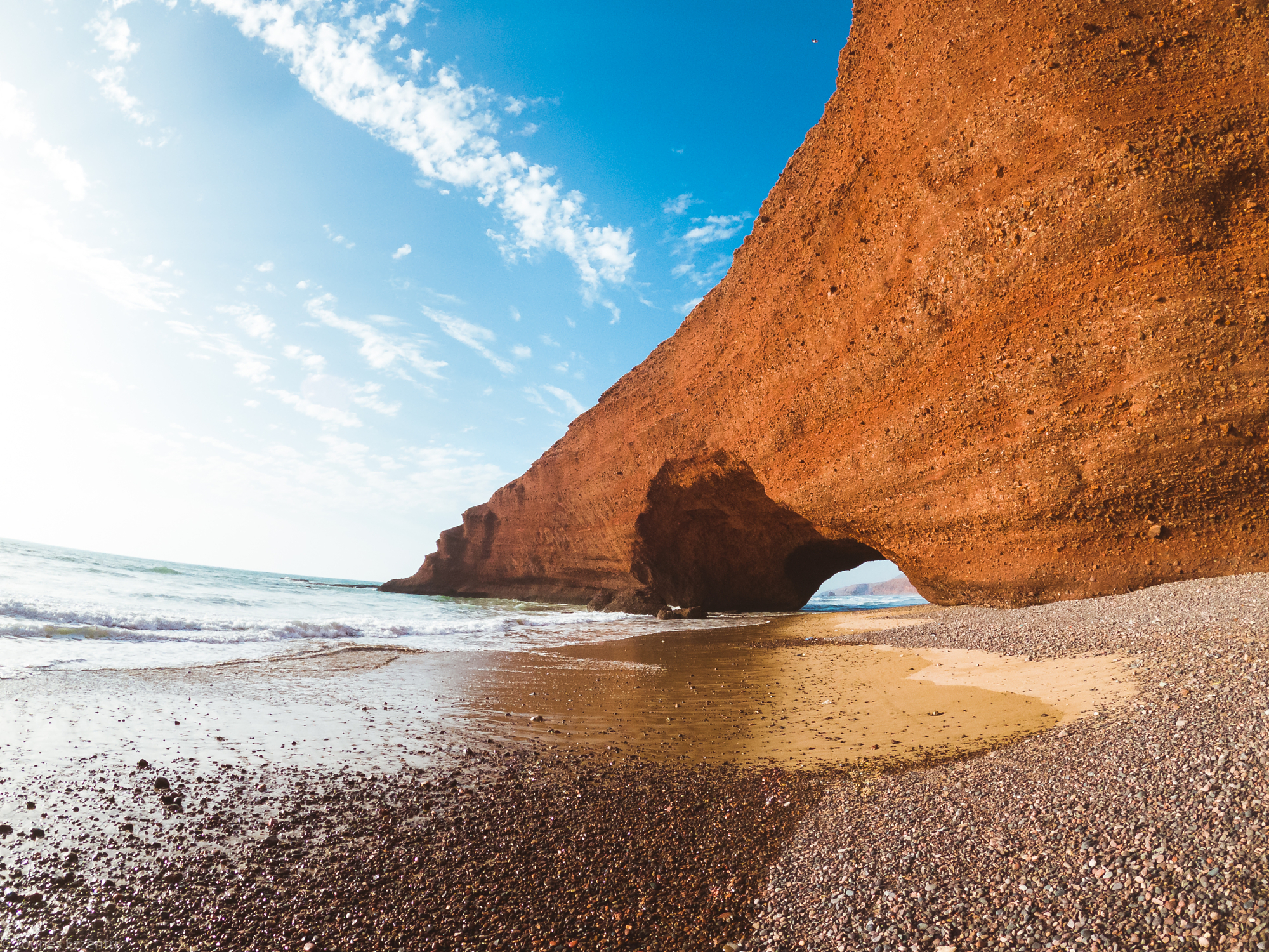 The massive natural red stone arch at Legzira Beach in Morocco curving over the ocean shoreline, a stunning location for adventure photography by Younes Ezzentri.