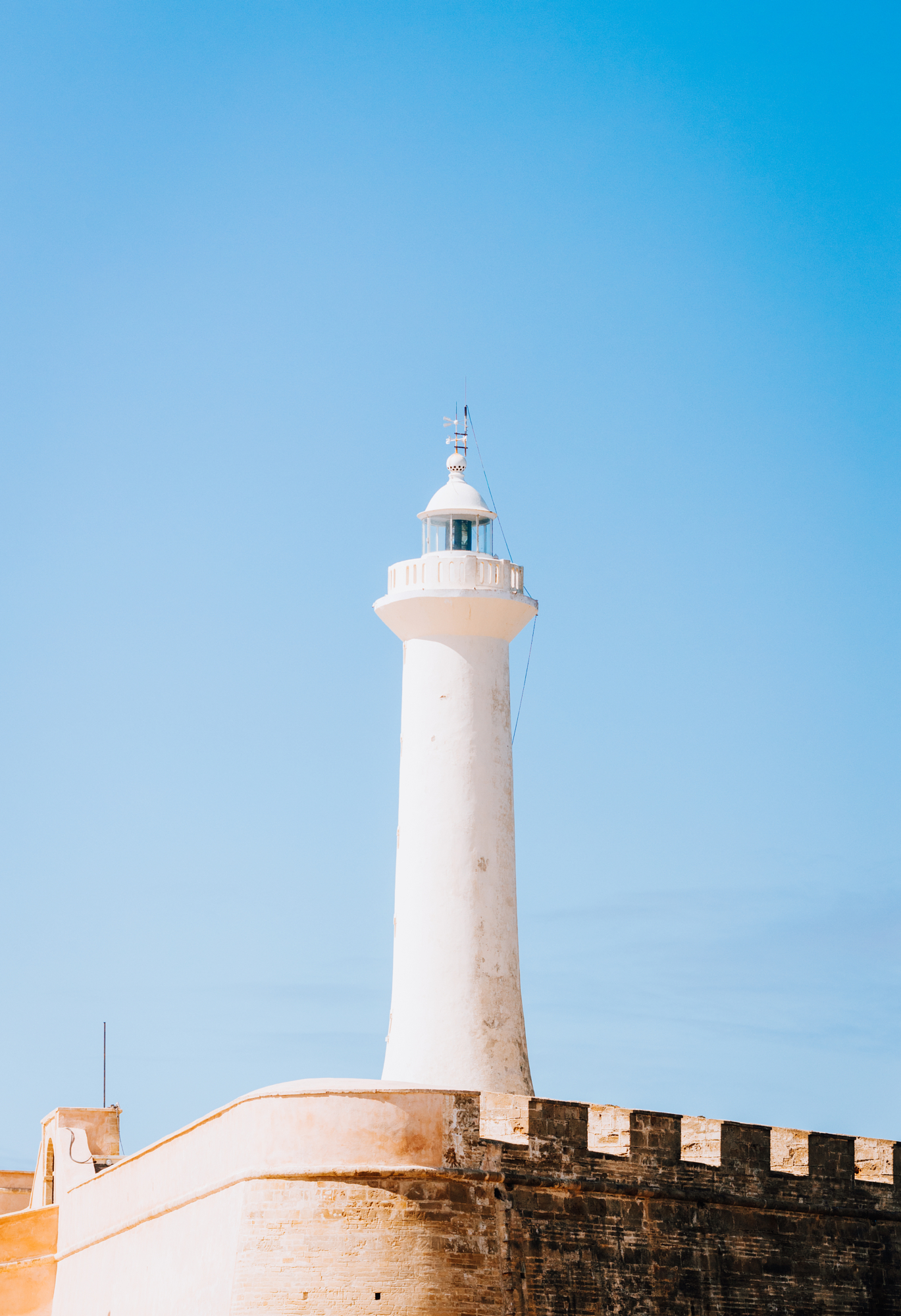 Low-angle view of a stark white lighthouse towering over defensive stone walls against a crisp blue sky, a clean and minimalist frame by Younes Ezzentri.