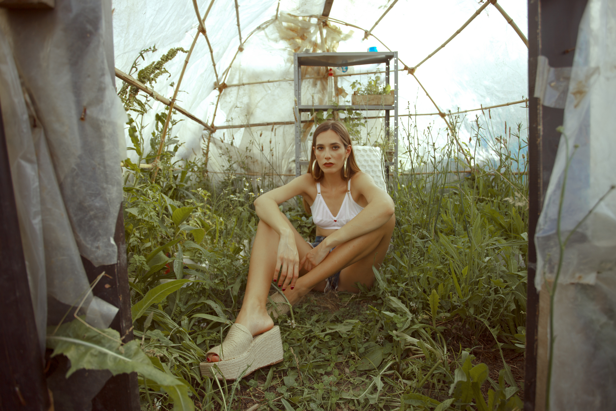 Environmental editorial fashion portrait featuring a model seated inside a greenhouse with natural green foliage.