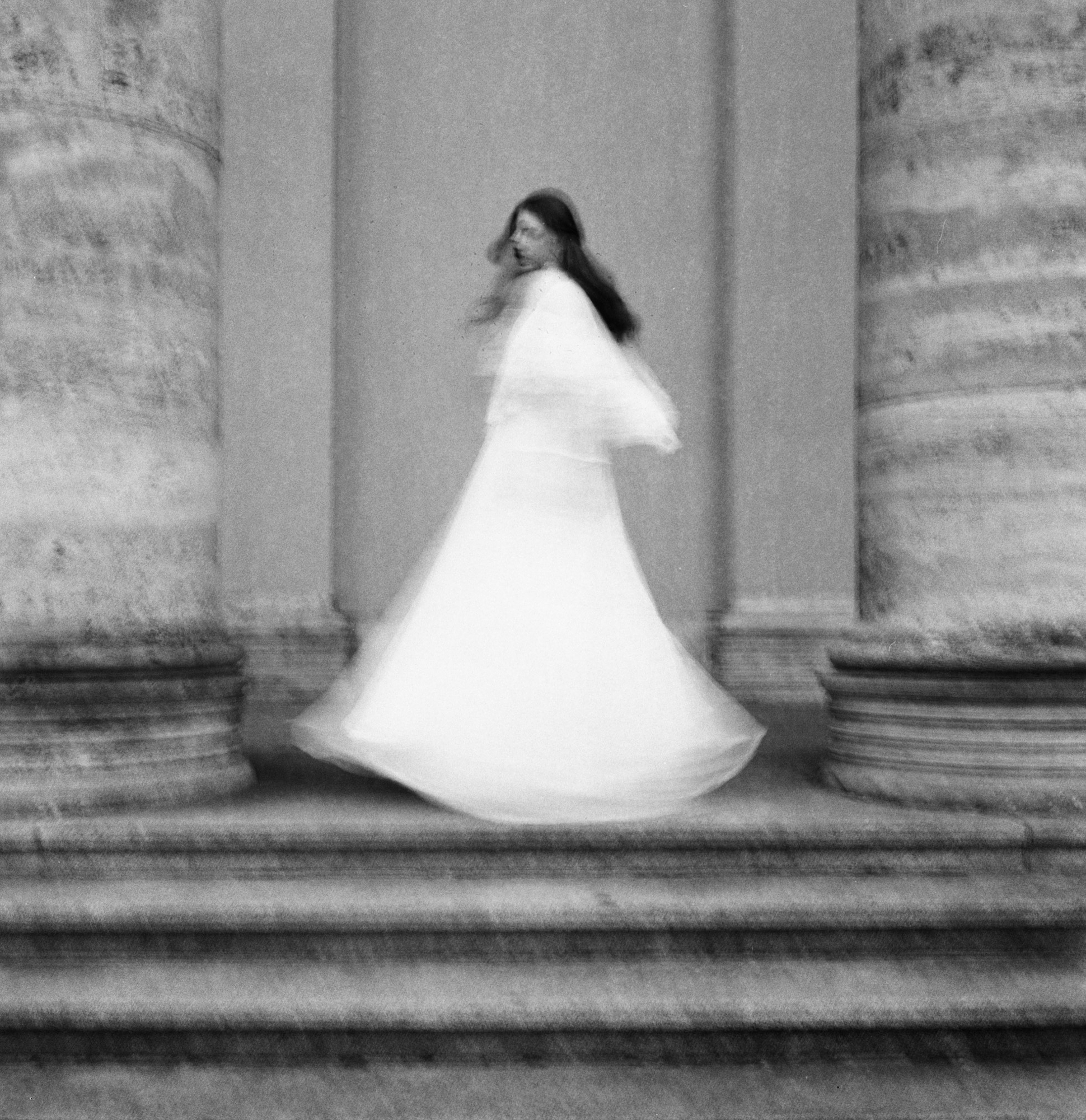 Fine art motion-blur black and white photograph by Holger Nitschke of a woman twirling in a white dress between classical stone columns.