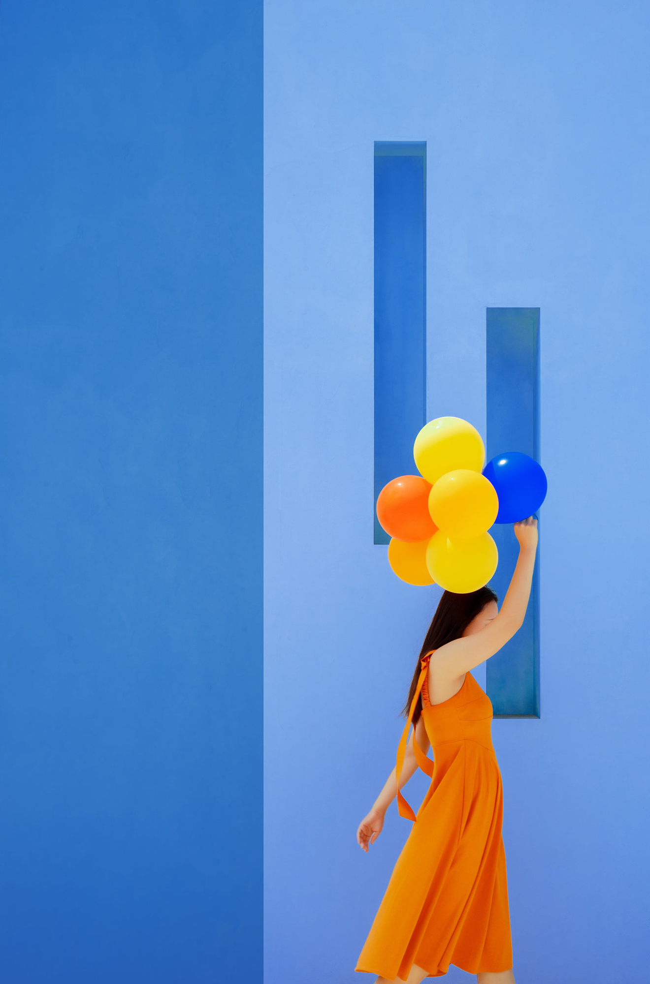 A woman in an orange dress carrying colorful balloons past a solid blue wall, showing how to capture vibrant summer colors cleanly.