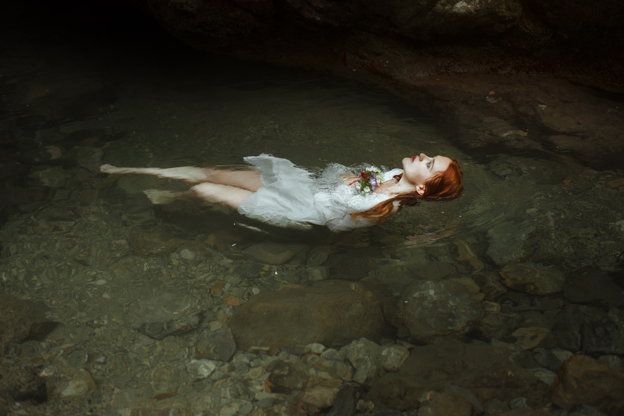 Romantic Ophelia-inspired fine art portrait of a red-haired woman floating in clear natural water with flowers.