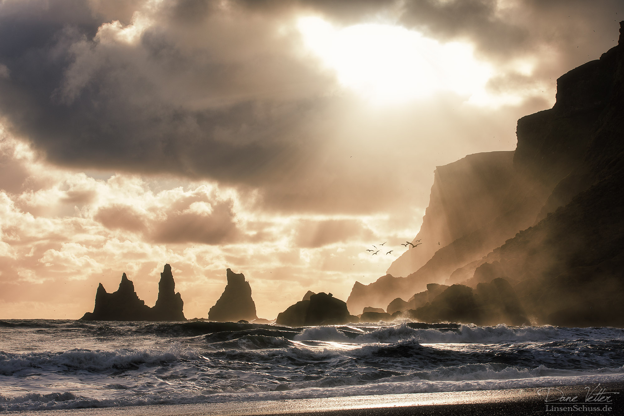 Dramatic landscape silhouette photography of rocky coastal sea stacks with sunlight breaking through dark clouds.
