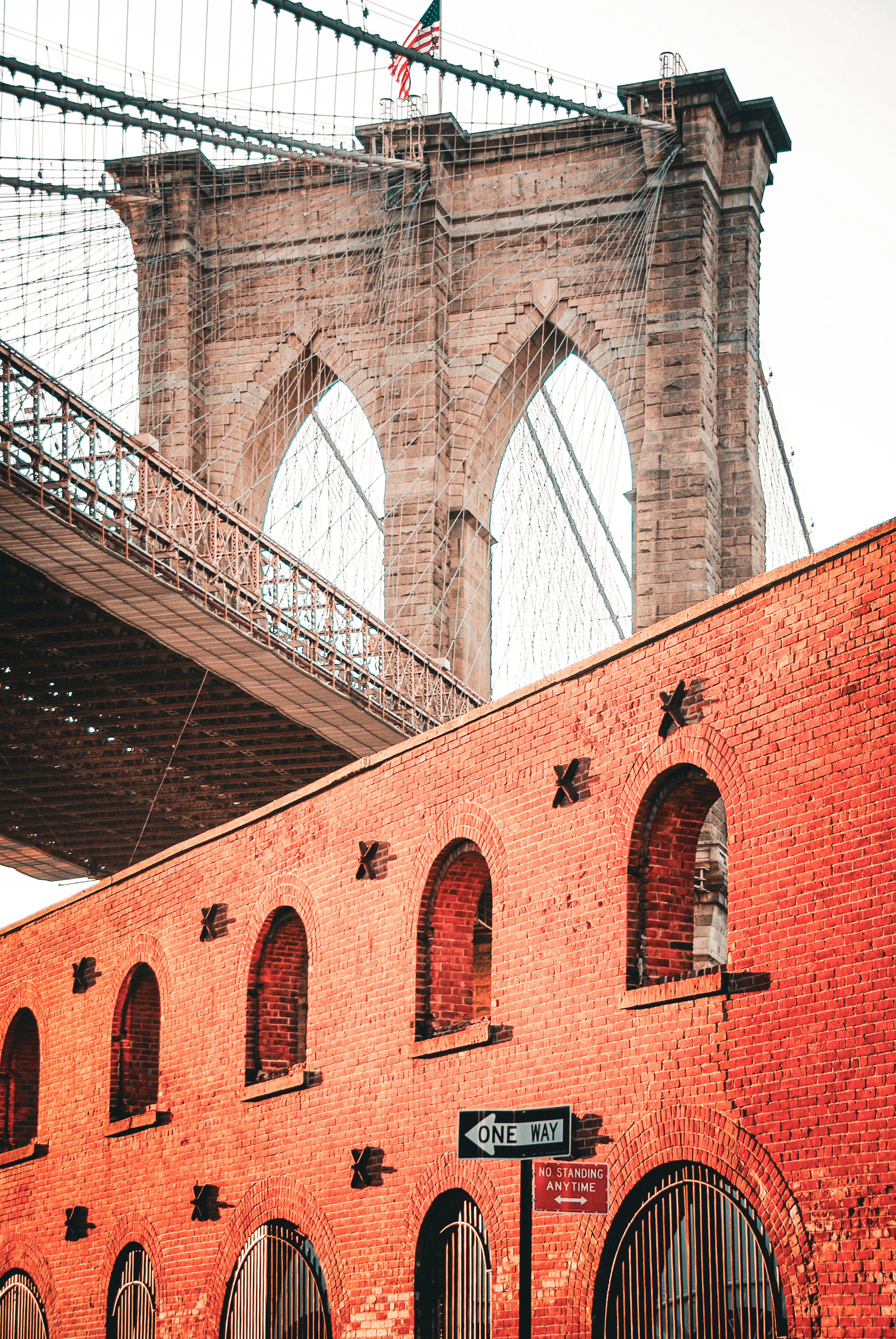 Brooklyn Bridge rising above a red brick warehouse wall and a one way sign