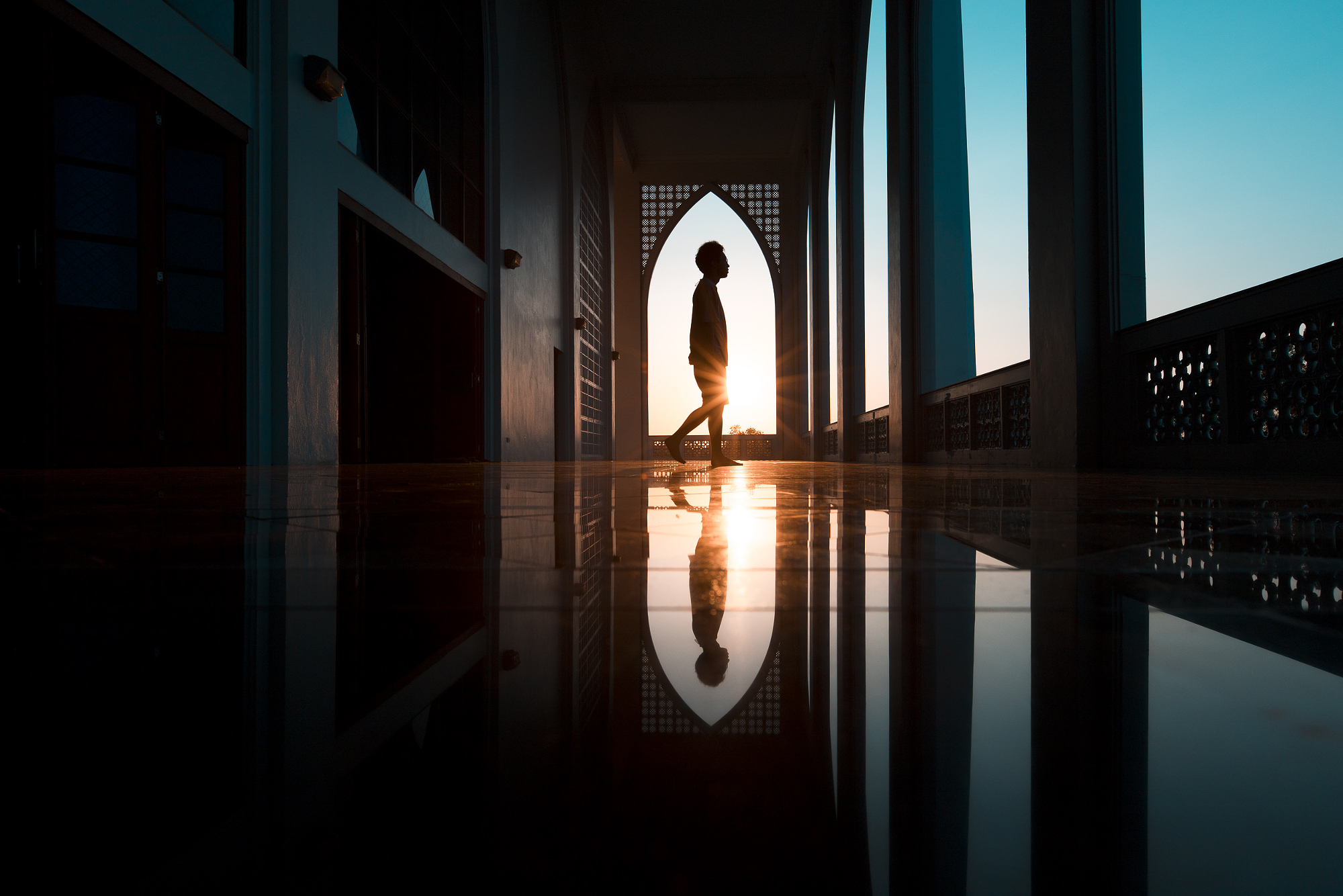 Silhouette walking through a mosque archway at sunset, warm golden light against a cool blue sky and reflective floor