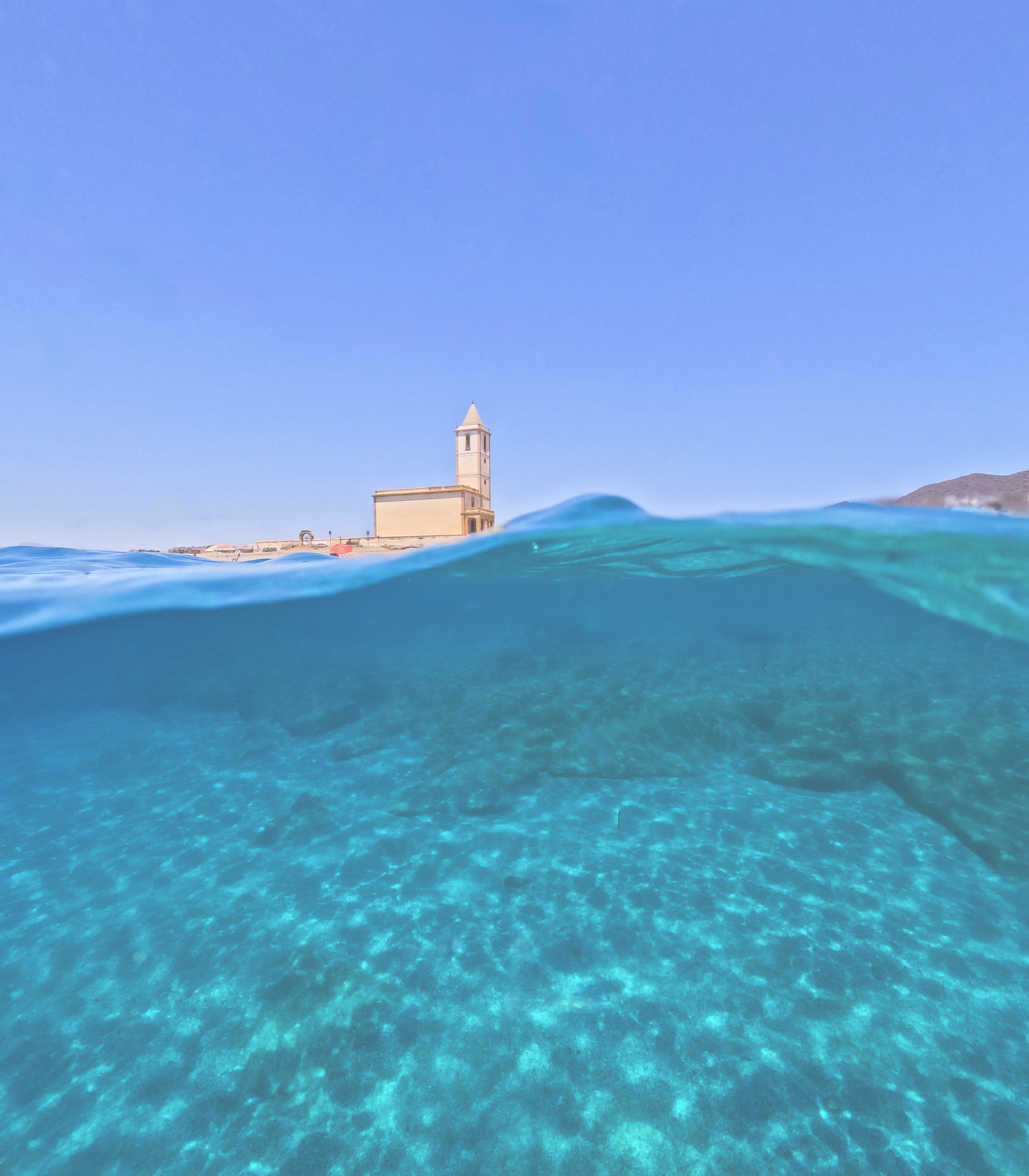Underwater split shot of clear turquoise sea water looking up at a historic coastal church in Spain by photographer Gonzalo Bendito
