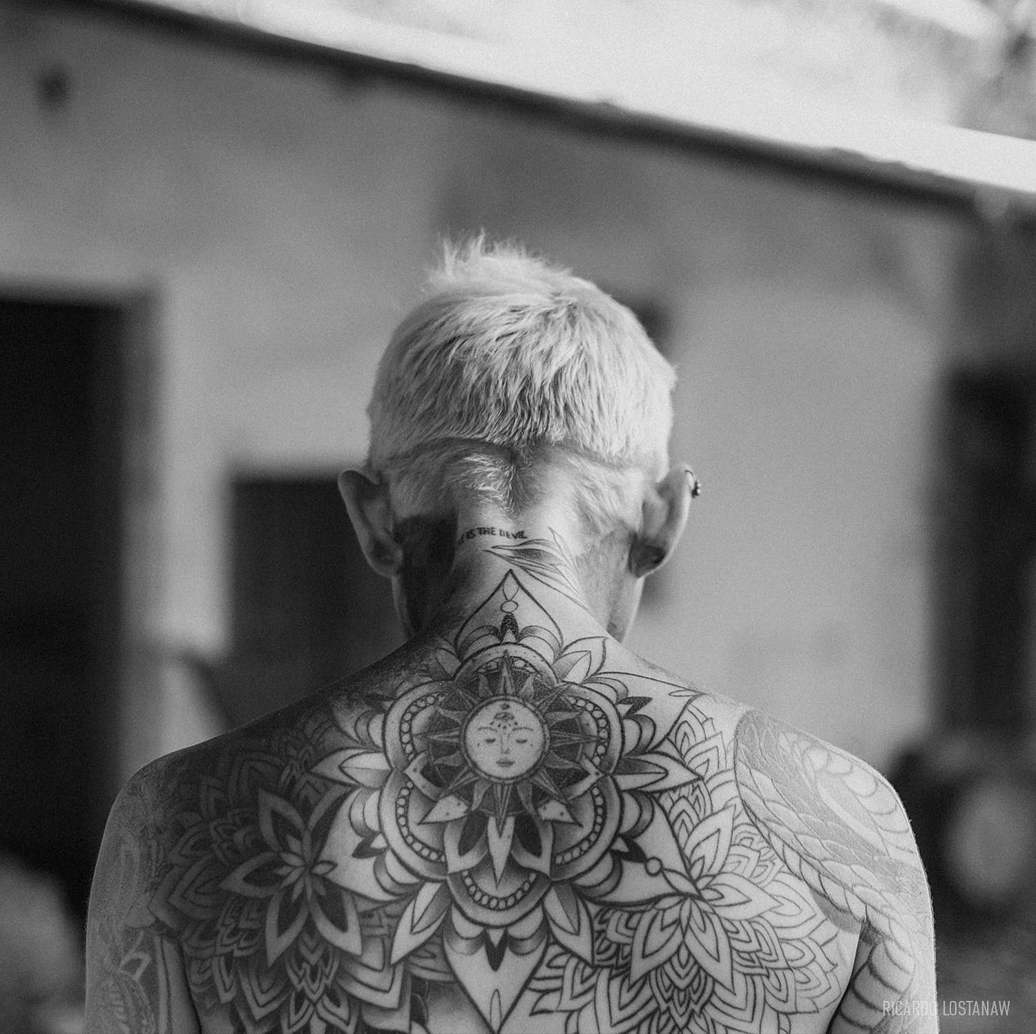 Black and white portrait from behind of a man with bleached hair and a large mandala tattoo across his back