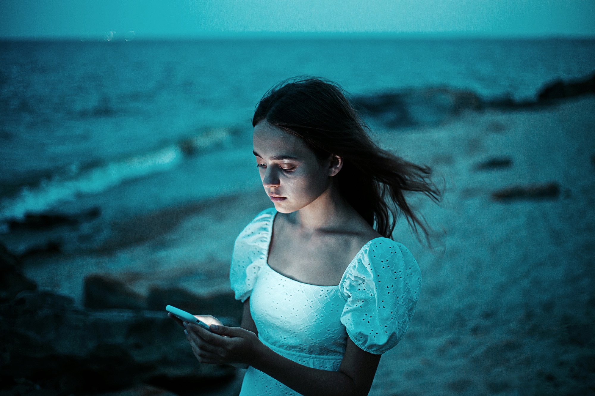 Girl in a white dress lit by her phone at blue hour, teal sea behind her — blue monochromatic color photography.