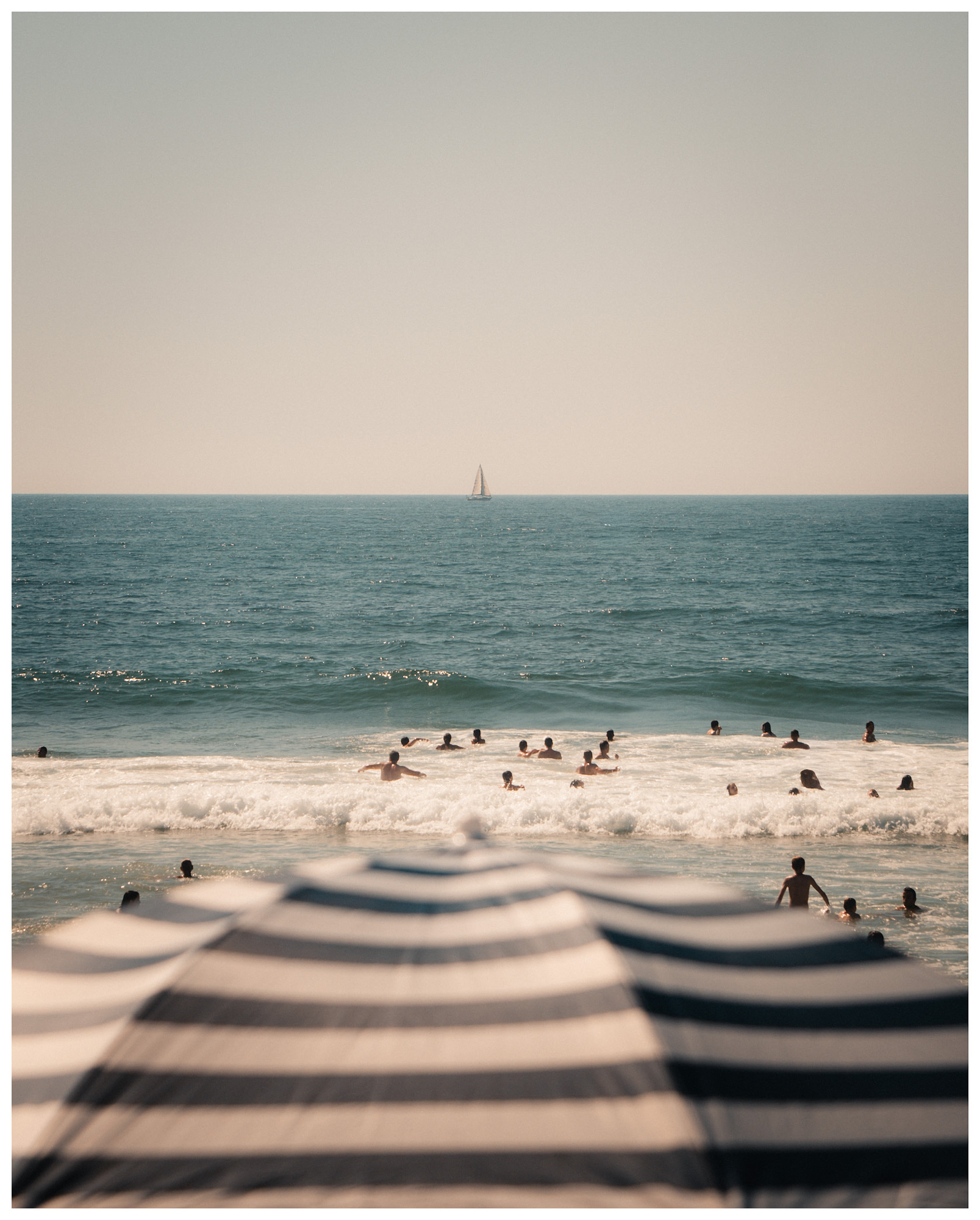 Swimmers in the surf seen past striped umbrellas, with a sailboat on the horizon