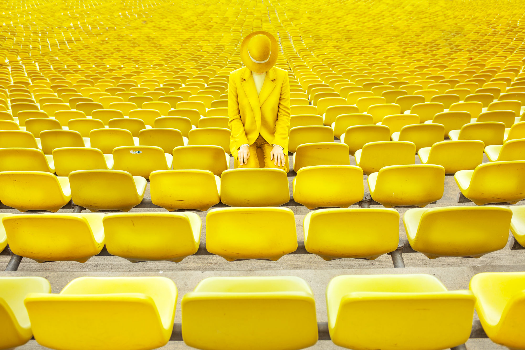 Person in a yellow suit and hat alone in rows of yellow stadium seats, a single color photography composition.