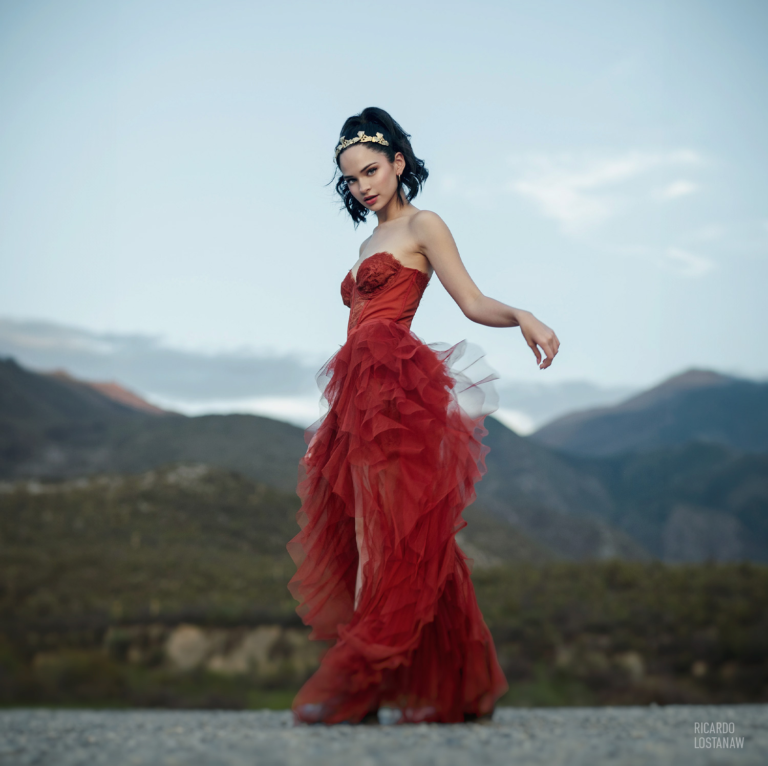 Young woman in a red tulle gown and gold headpiece against a mountain backdrop