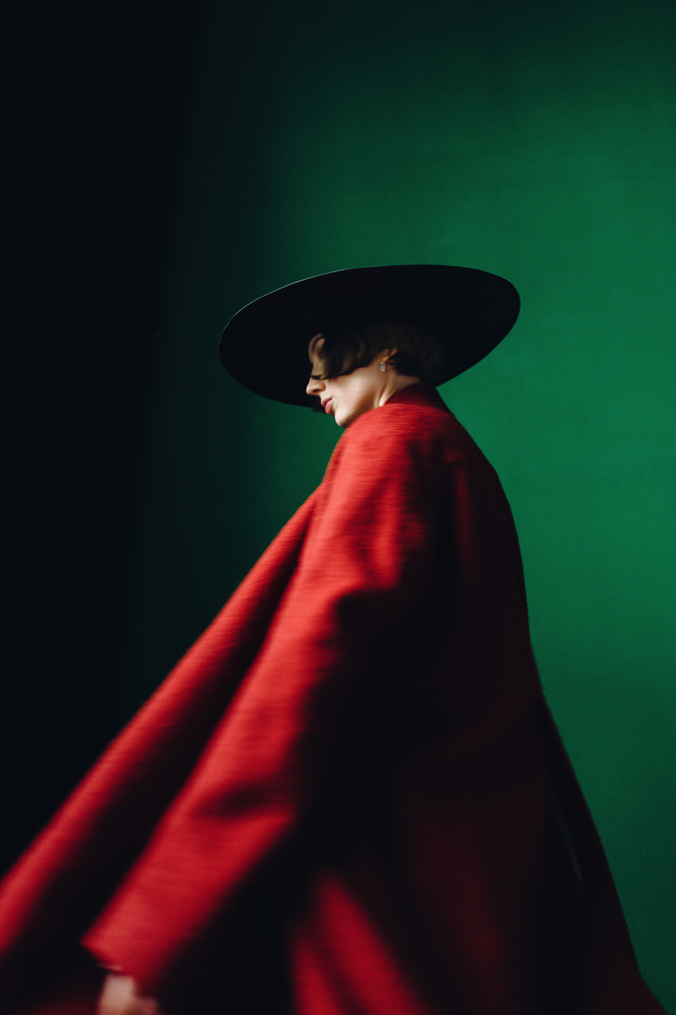 Woman in a flowing red coat and wide-brimmed hat photographed against a deep green backdrop