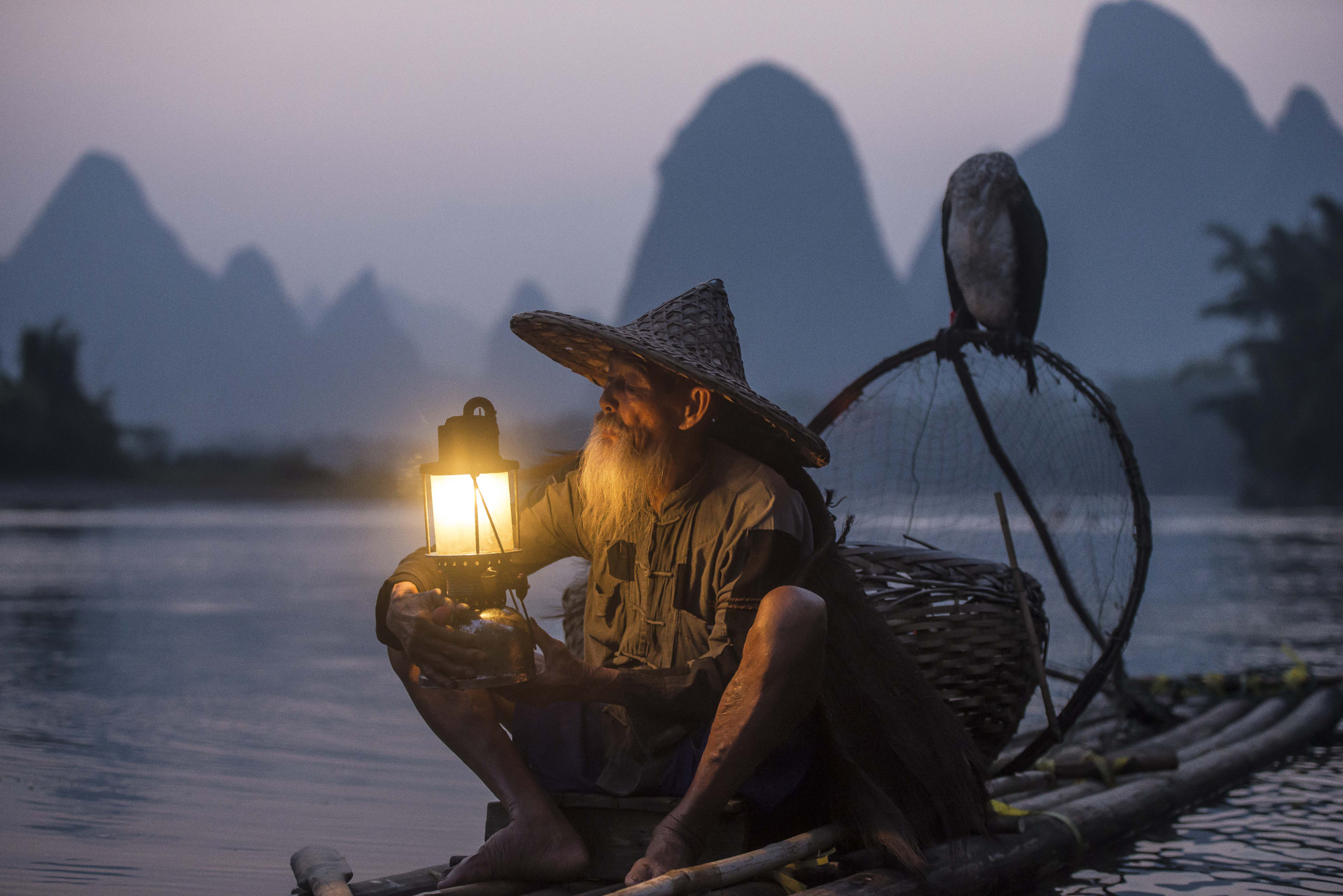 Traditional cormorant fisherman holding a glowing lantern on a river raft in China, Gonzalo Bendito photograph